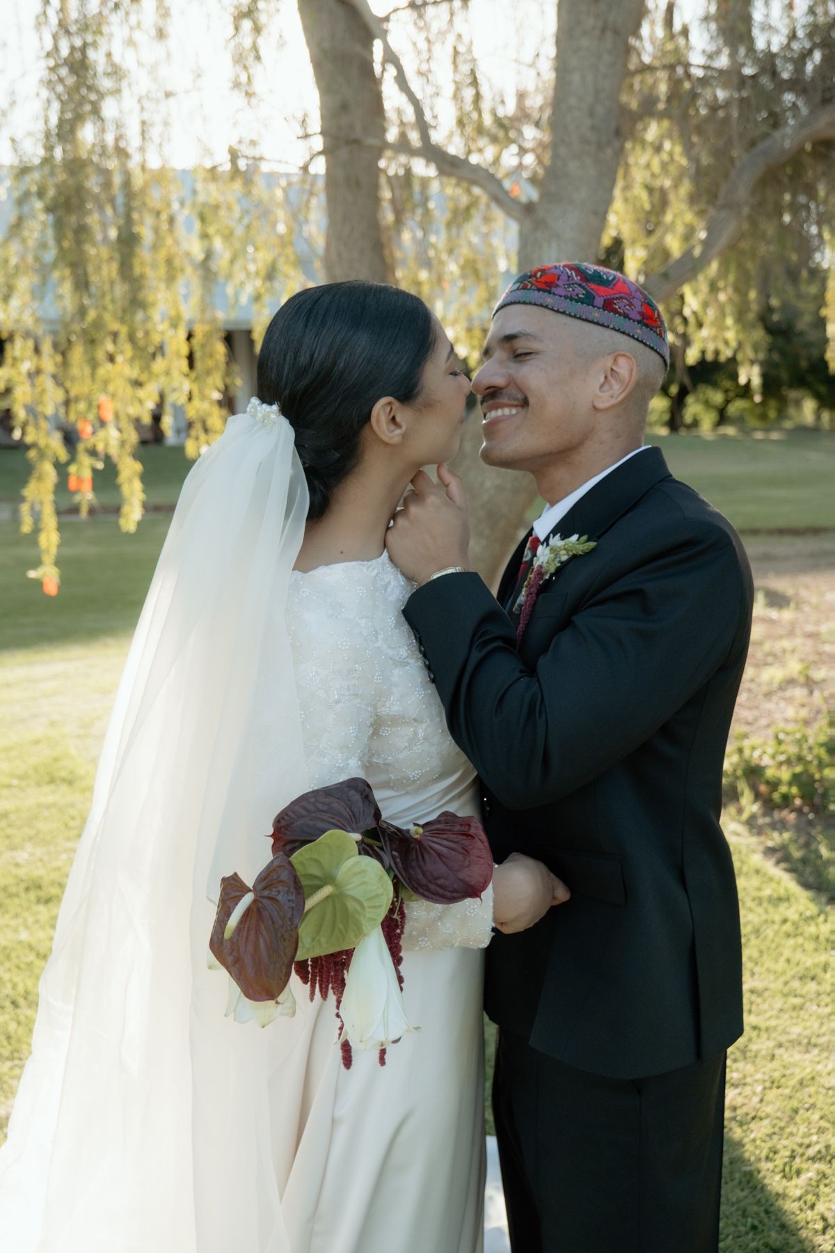 Bride in white gown with veil kissing groom wearing traditional embroidered kufi cap outdoors.