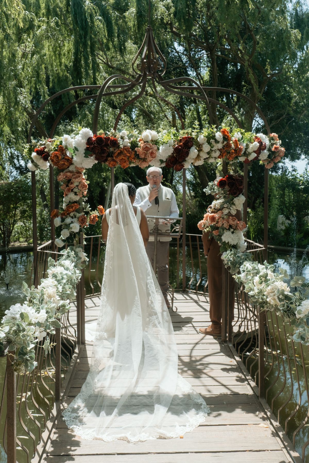 Bride in flowing veil walks toward officiant on floral-lined bridge at elegant outdoor garden wedding.