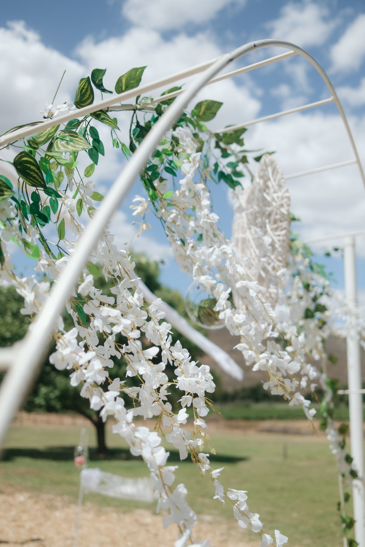 White floral wedding arch adorned with cascading wisteria blooms and green leaves against a bright blue sky.