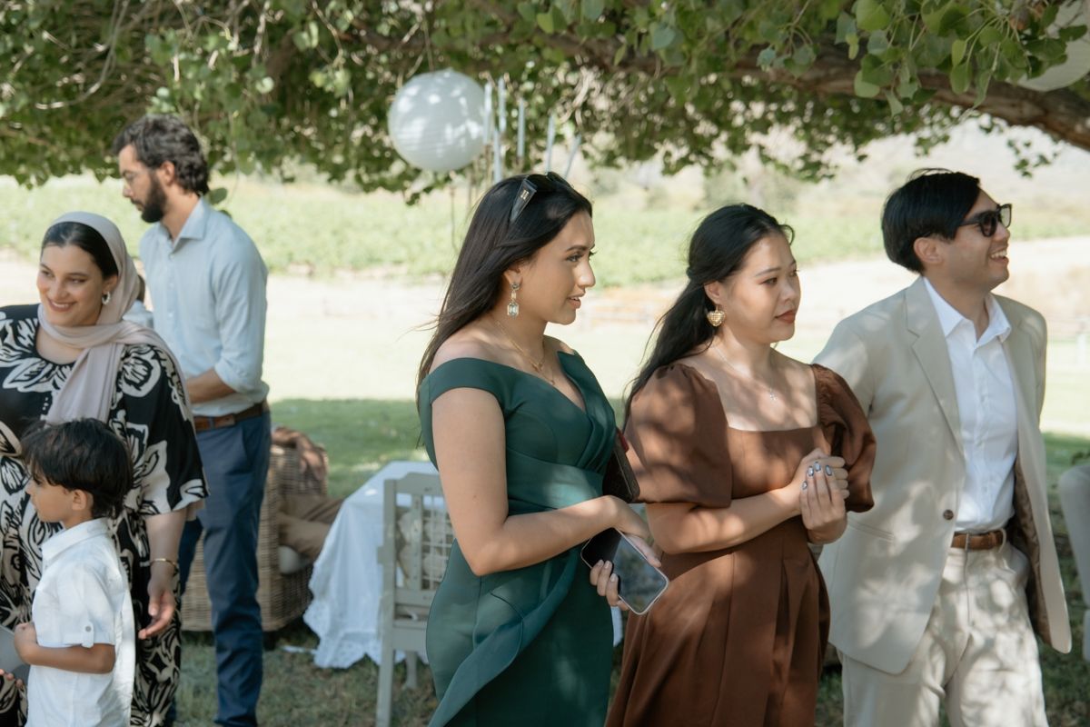 Wedding guests mingle outdoors under trees at elegant garden reception celebration.