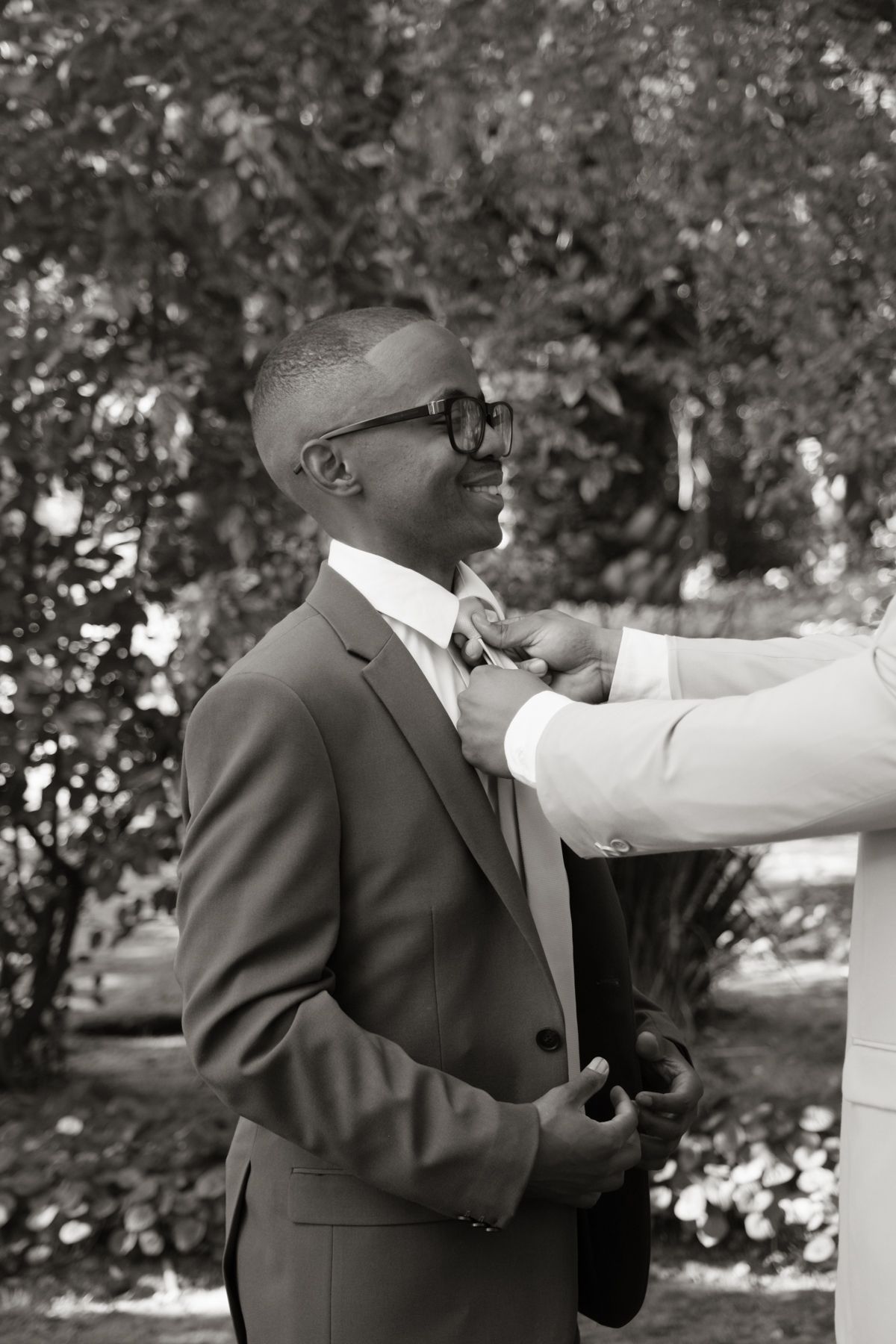 Black and white photo of smiling groom in suit having boutonniere pinned on lapel outdoors.