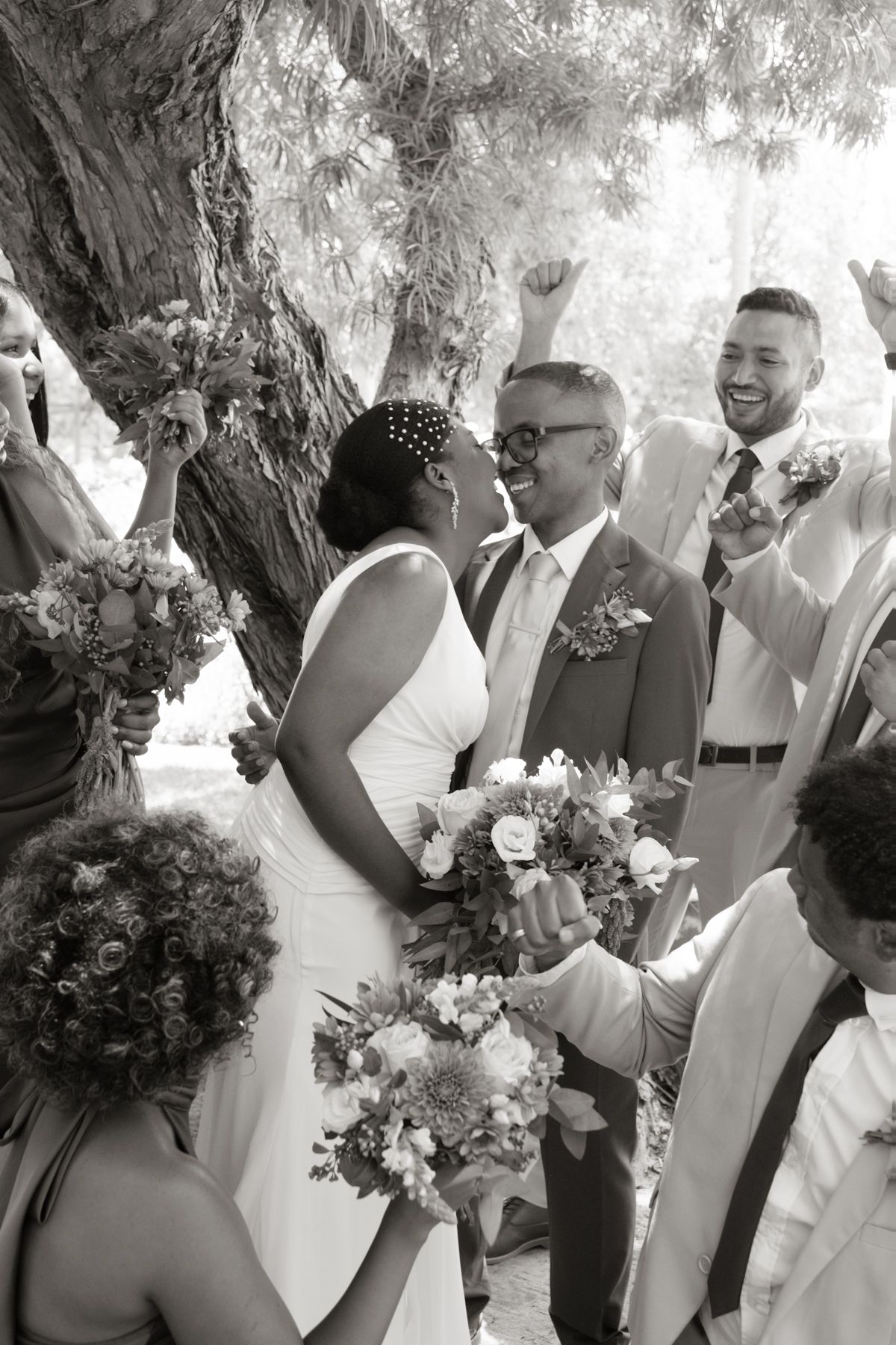 Black and white wedding photo of bride and groom kissing surrounded by joyful wedding party under a tree.