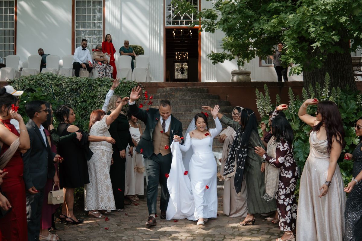 Joyful newlyweds exit ceremony as guests celebrate with raised hands outside historic venue.