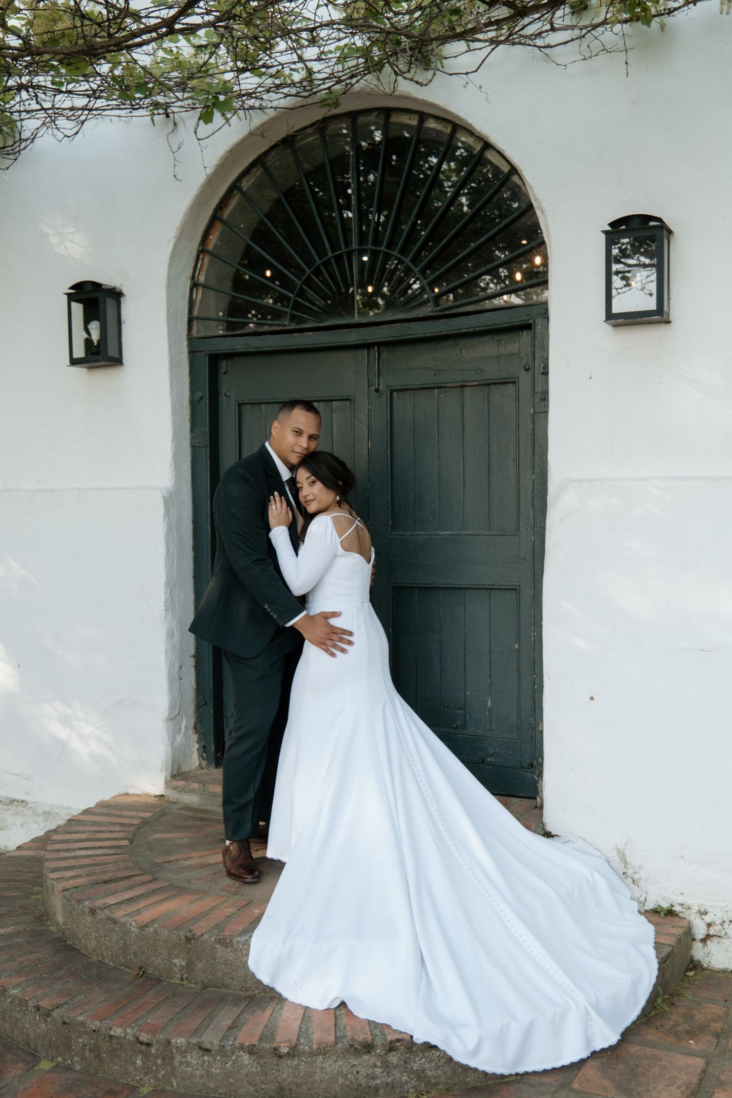 Bride and groom share romantic embrace before ornate green arched door, her white mermaid gown trailing on brick steps.