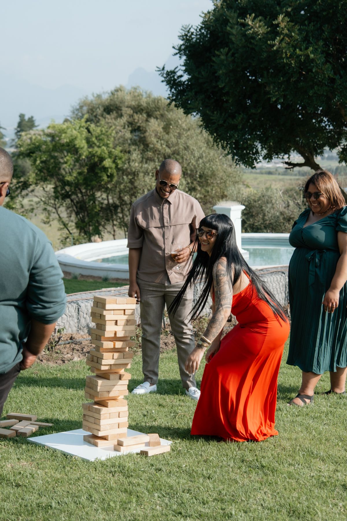 Wedding guests playing giant Jenga lawn game outdoors near pool on sunny celebration day.