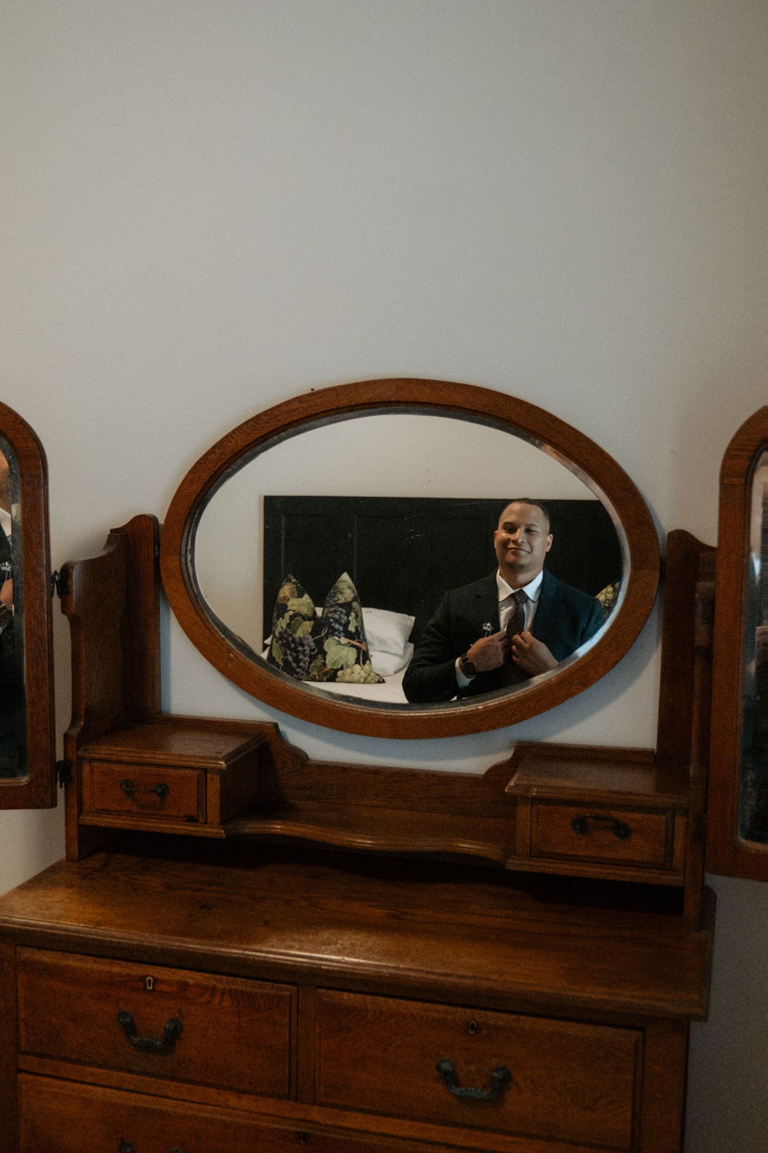 Groom adjusting his tie reflected in an antique oval wooden dresser mirror on wedding day.