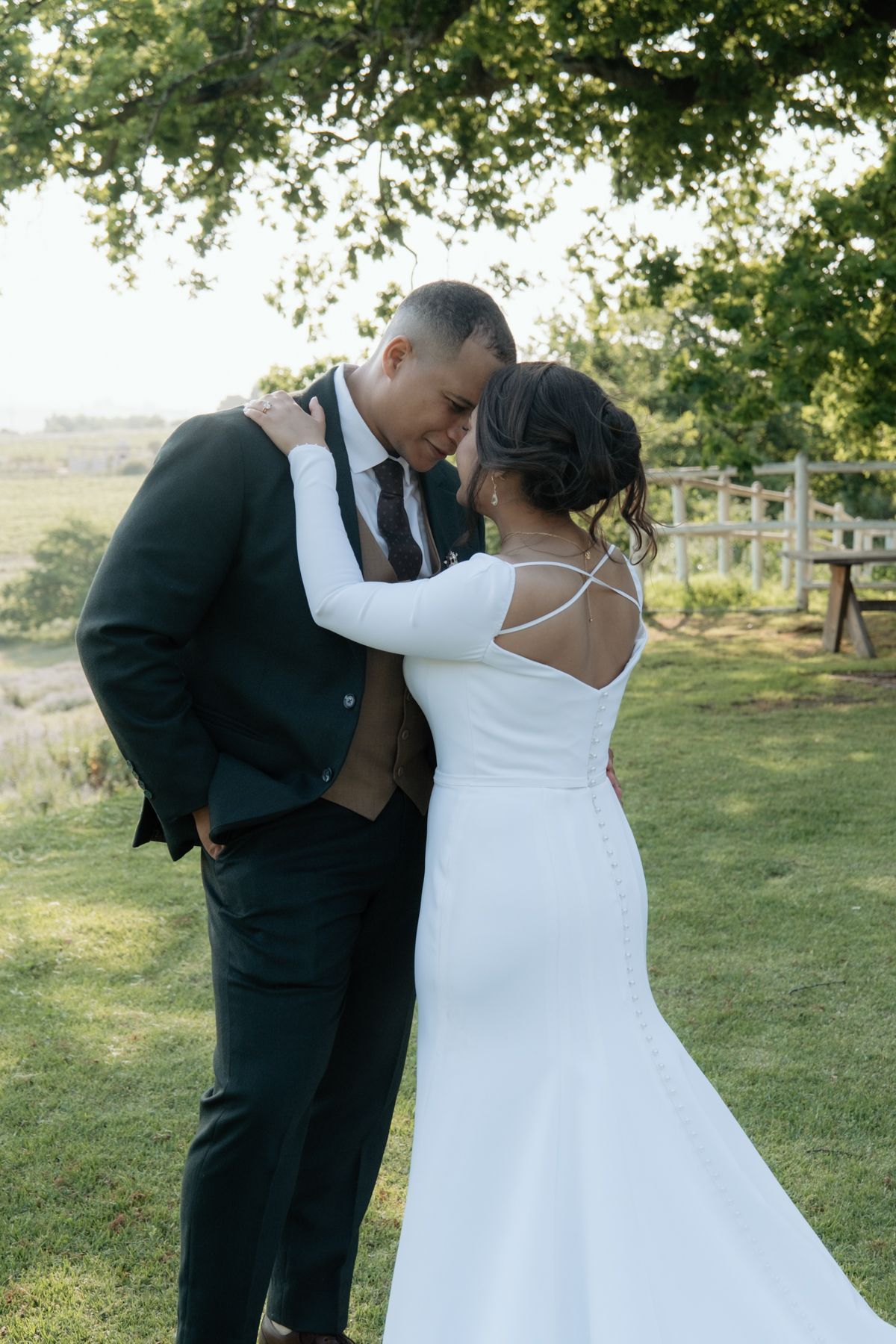 A bride and groom share an intimate moment embracing under a large tree in a lush green outdoor setting.