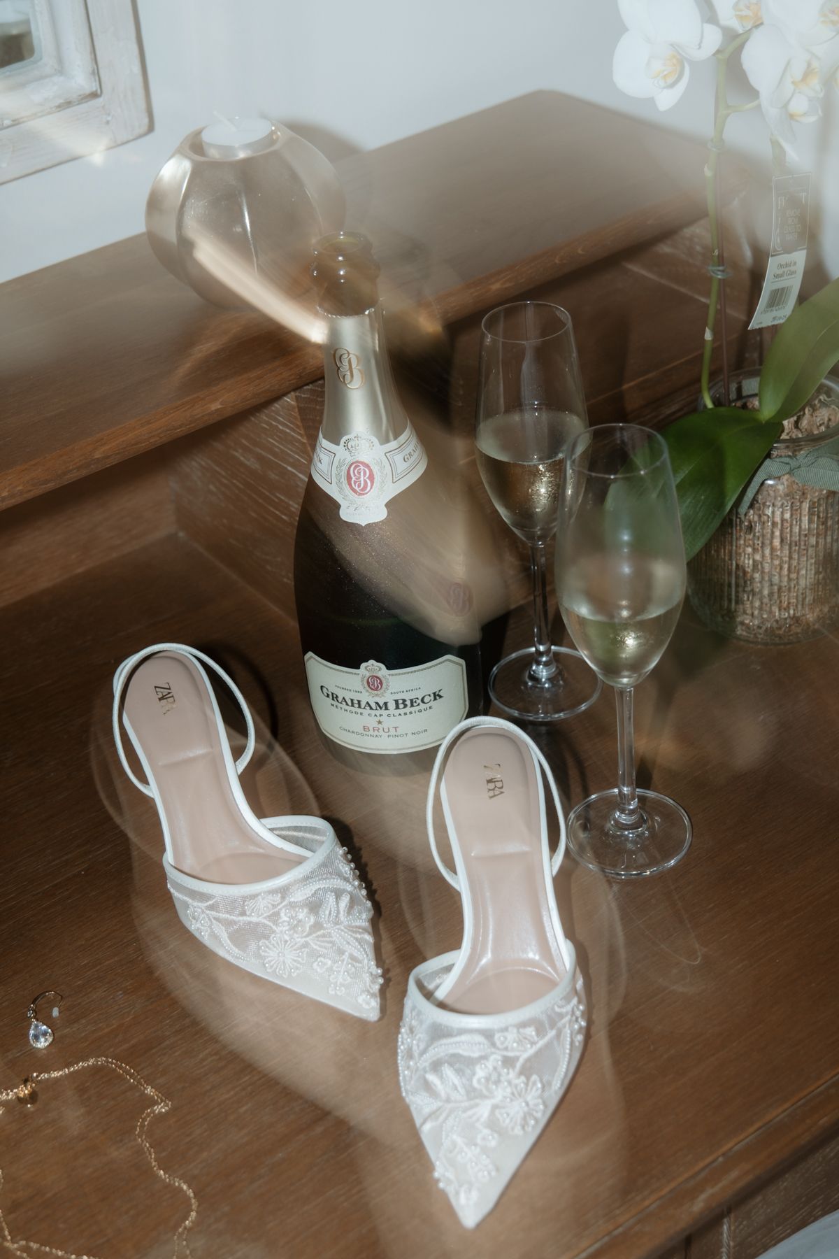 Elegant flat lay of white lace bridal shoes beside Graham Beck champagne and glasses on wooden dresser.
