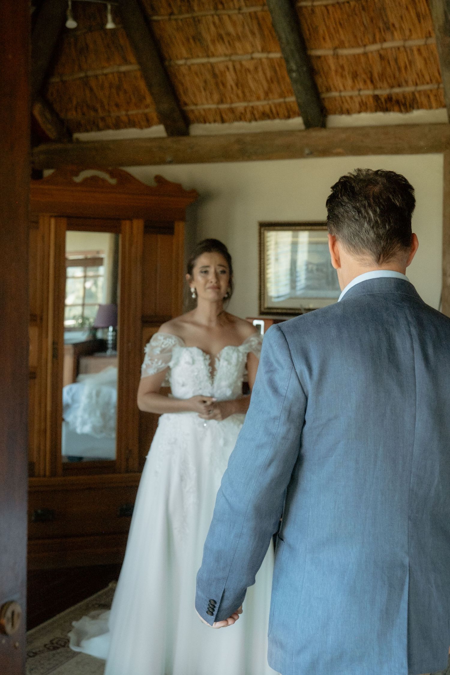 Groom sees bride in lace off-shoulder wedding dress for the first time in a rustic thatched room.