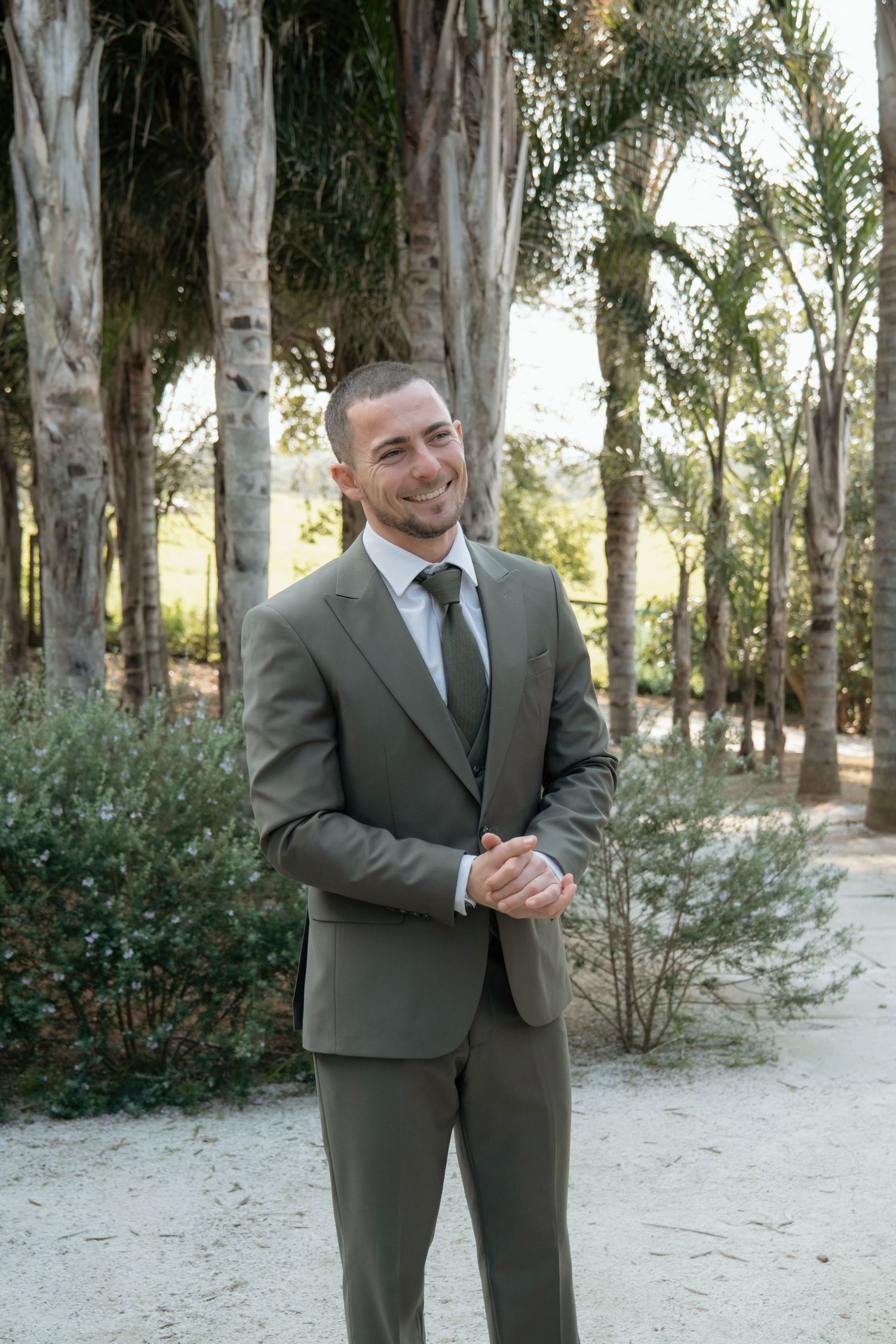 Smiling groom in olive green suit standing outdoors among tall trees on wedding day.