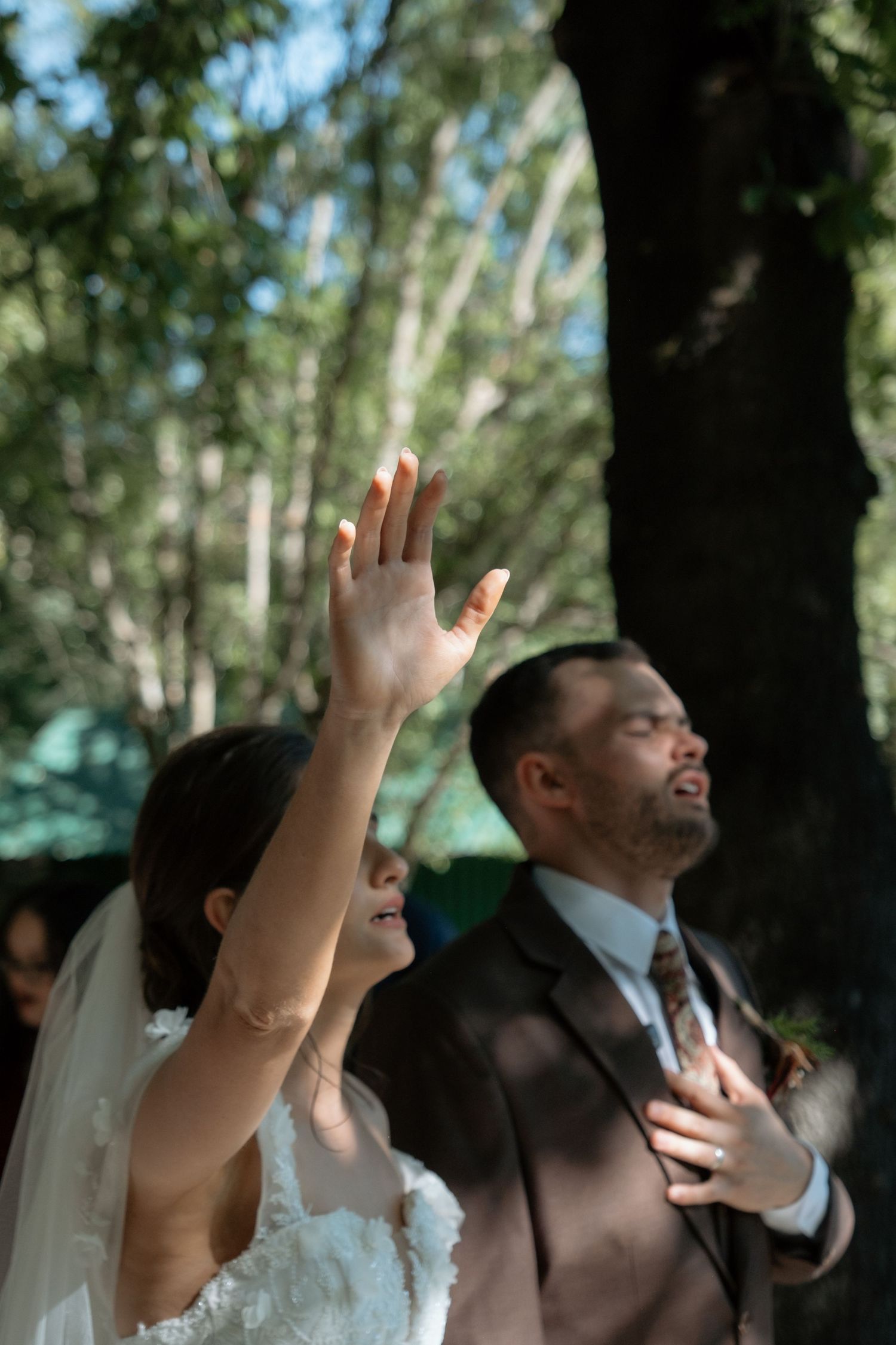 Bride raising her hand during outdoor wedding ceremony vows while groom stands beside her under trees.