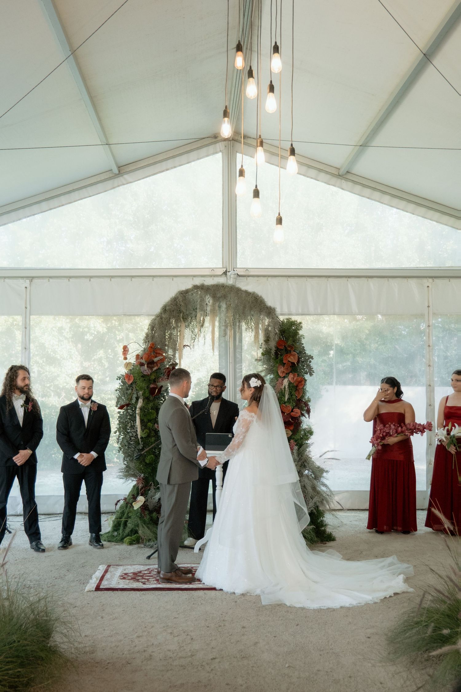 Bride and groom exchanging vows under floral arch in elegant tent wedding ceremony.
