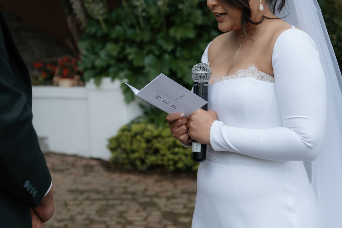 Bride in white long-sleeve gown reading vows from a booklet during outdoor wedding ceremony.
