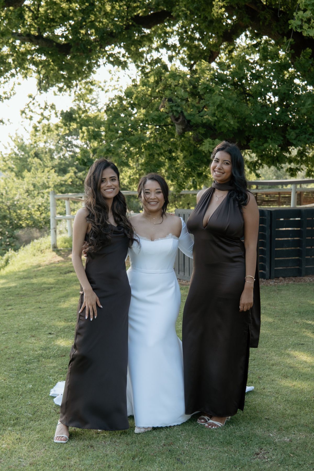 Bride in strapless white gown poses with two bridesmaids in dark brown dresses on green lawn.