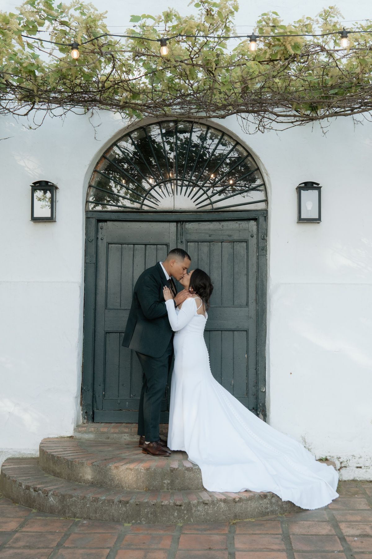 Bride and groom share a romantic kiss on steps before an arched wooden door with string lights above.