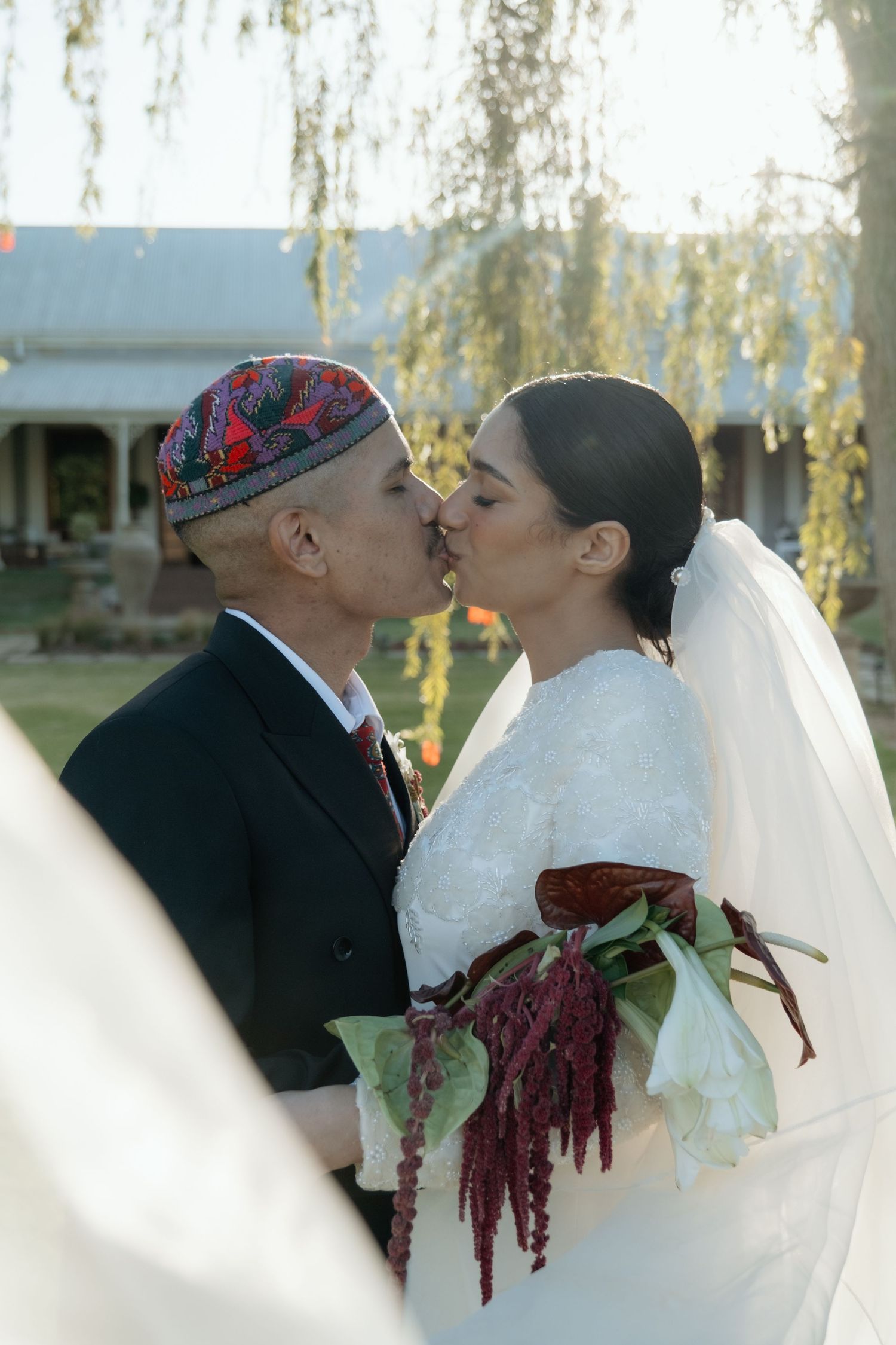 Bride and groom share a kiss outdoors, groom wearing embroidered skullcap, bride holding red and white bouquet.