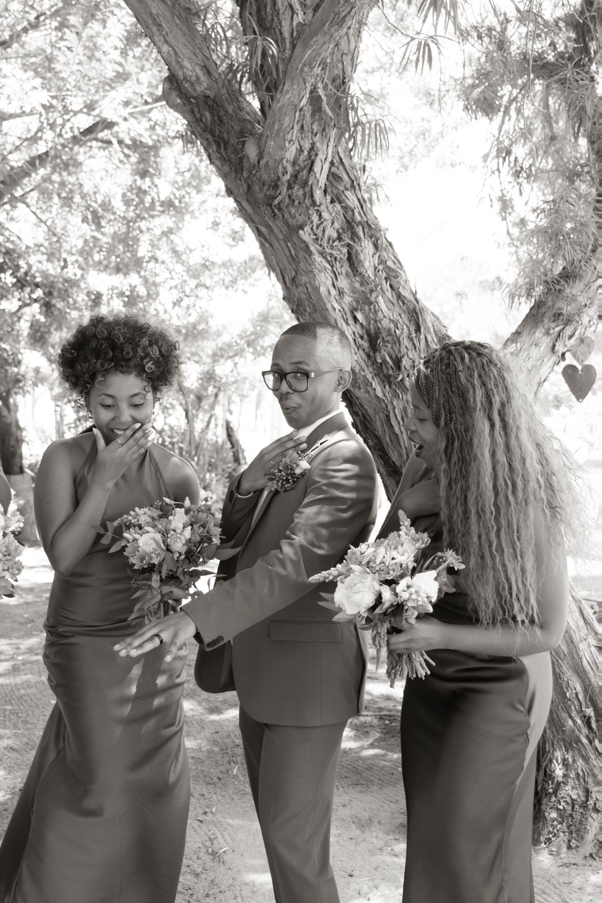 Black and white photo of groom in suit sharing a fun moment with two bridesmaids holding bouquets under a tree.