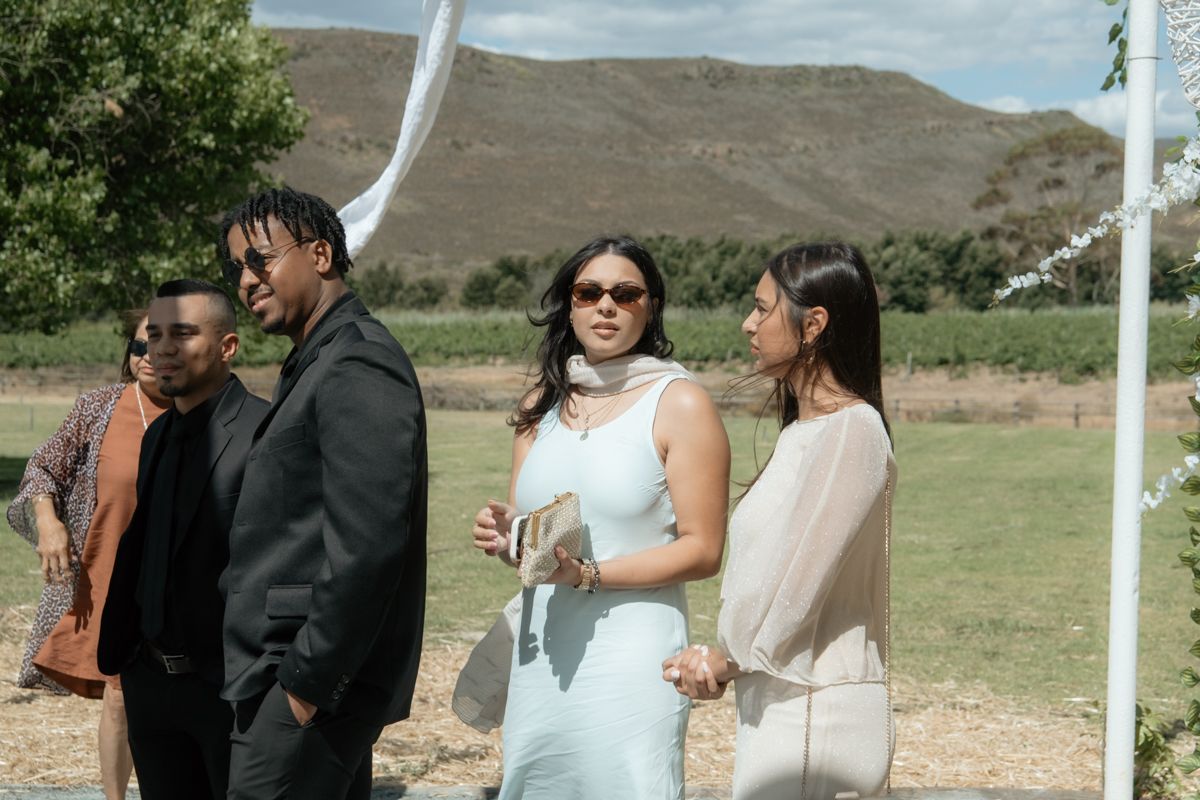 Wedding guests in formal attire socializing outdoors near a white arch with mountain views in the background.
