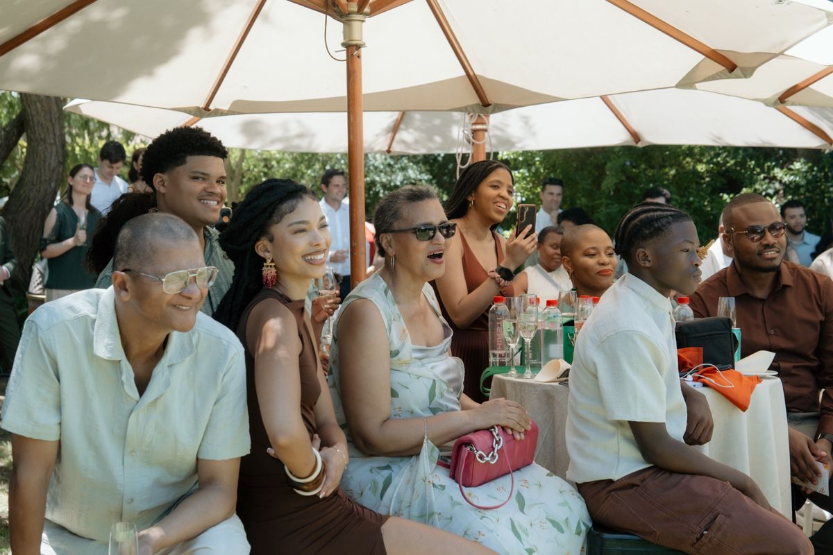 Diverse group of joyful wedding guests laughing and socializing under patio umbrella at outdoor reception.