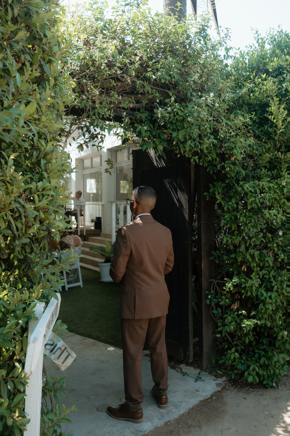Groom in brown suit stands at ivy-covered garden archway entrance at outdoor wedding venue.