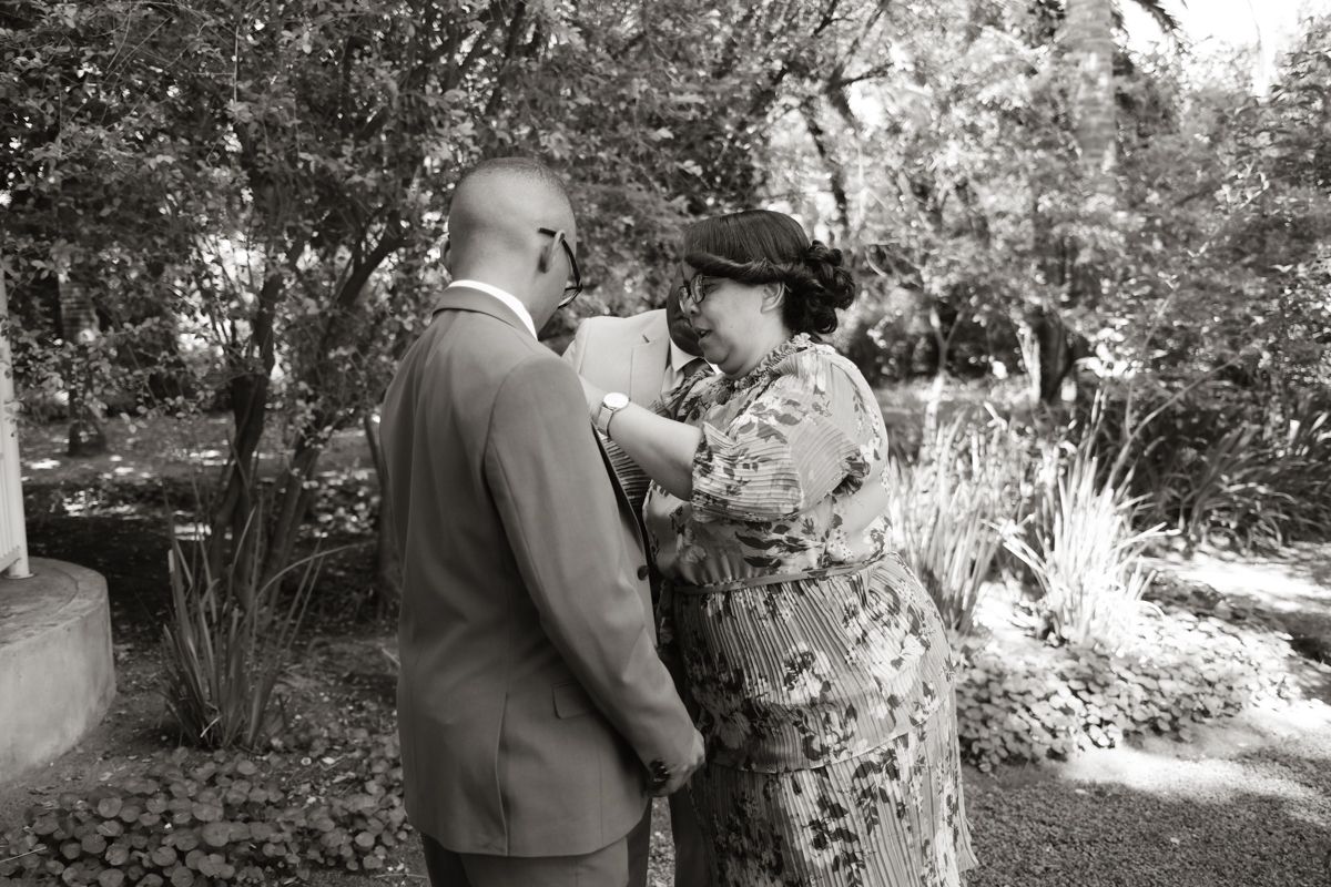 Black and white photo of groom having boutonniere pinned by woman in floral dress in garden.