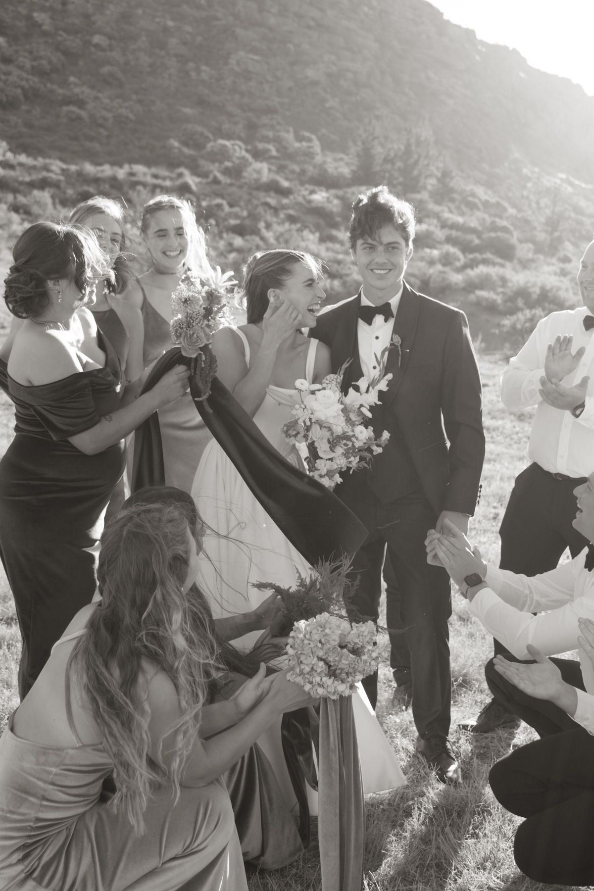 Black and white photo of a bride and groom laughing with bridesmaids during an outdoor wedding celebration.