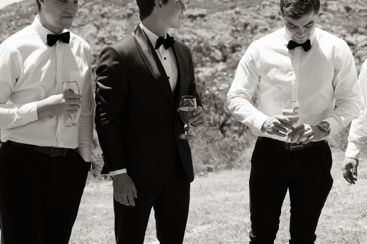 Three groomsmen in black tuxedos and bow ties holding drinks outdoors in a black and white wedding photo.