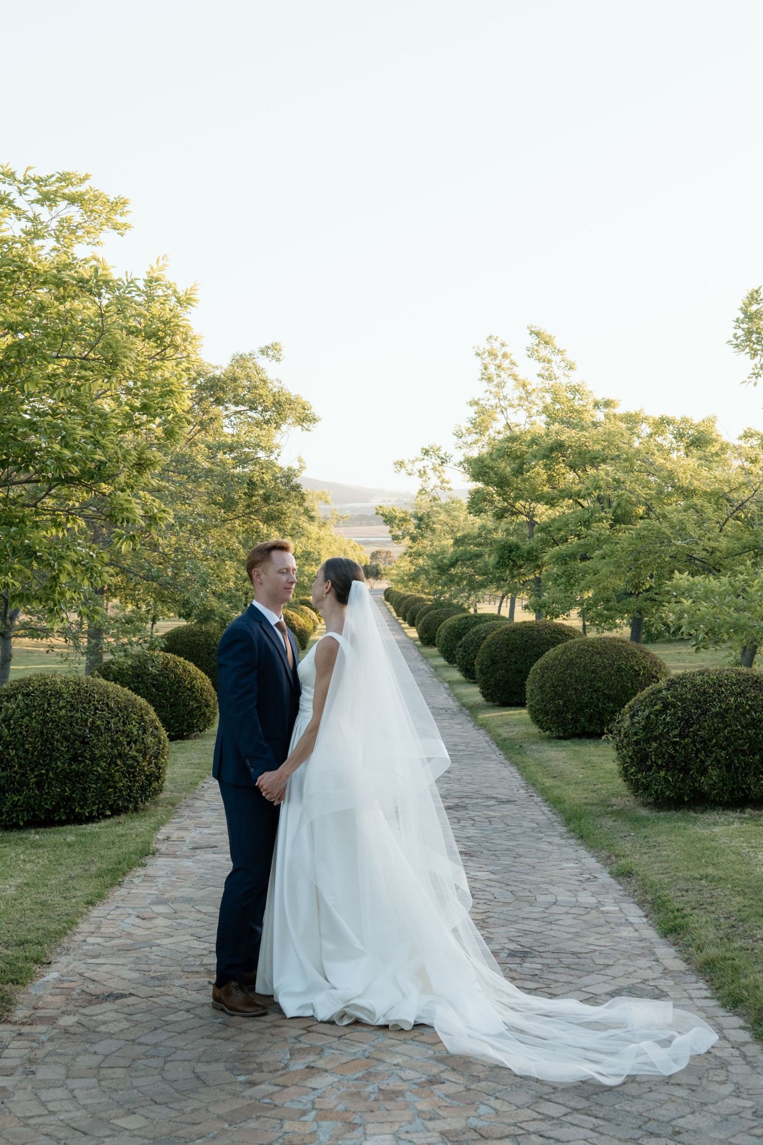 Bride and groom hold hands on a tree-lined garden path, bride in flowing white gown with long veil trailing behind.