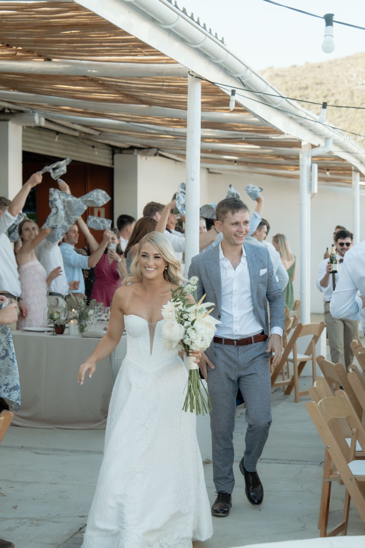 Smiling bride in strapless gown and groom in grey suit walk through cheering guests at outdoor wedding reception.
