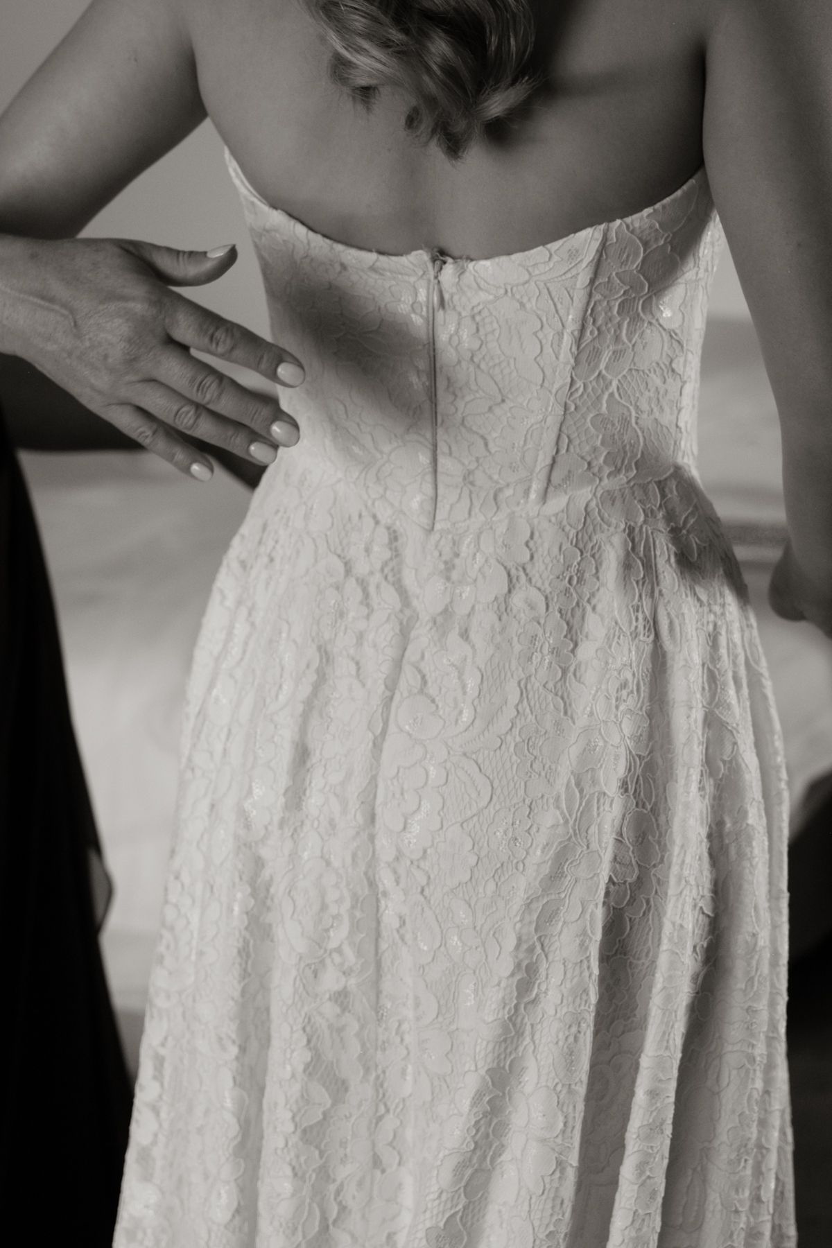 Black and white photo of a bride having her lace wedding dress zipped up from behind.