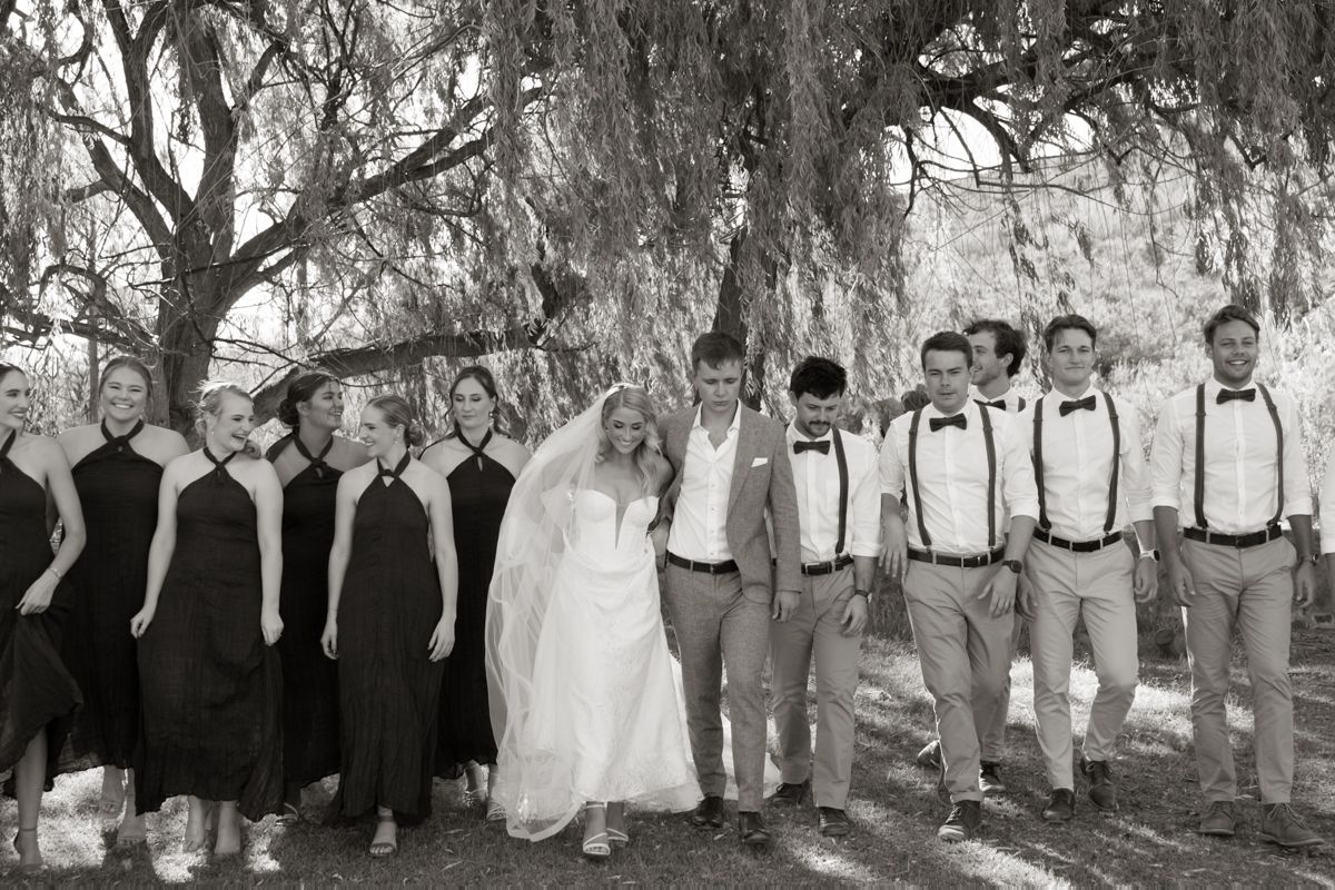 Black and white photo of wedding party posing together under a large willow tree.