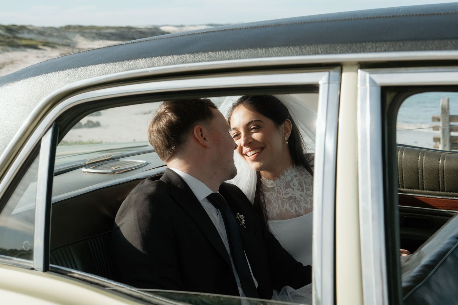 Bride and groom sharing a romantic moment inside a vintage classic car on their wedding day.