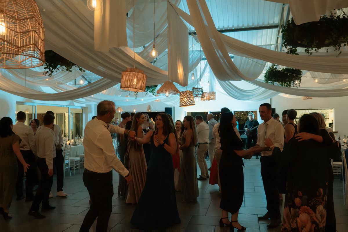 Wedding guests dance under white draped ceiling with warm Edison bulb lighting at elegant reception.