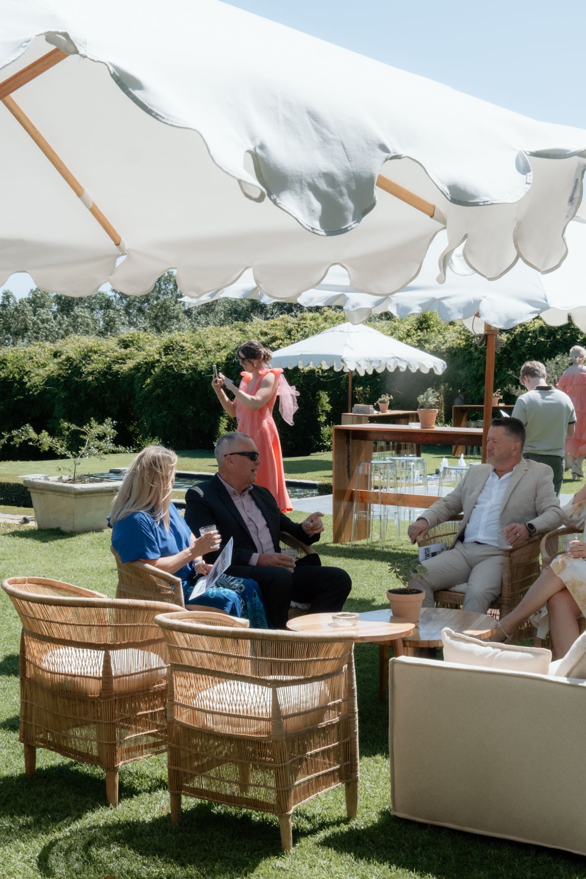 Wedding guests relax in wicker chairs under a white scalloped tent in sunny garden.