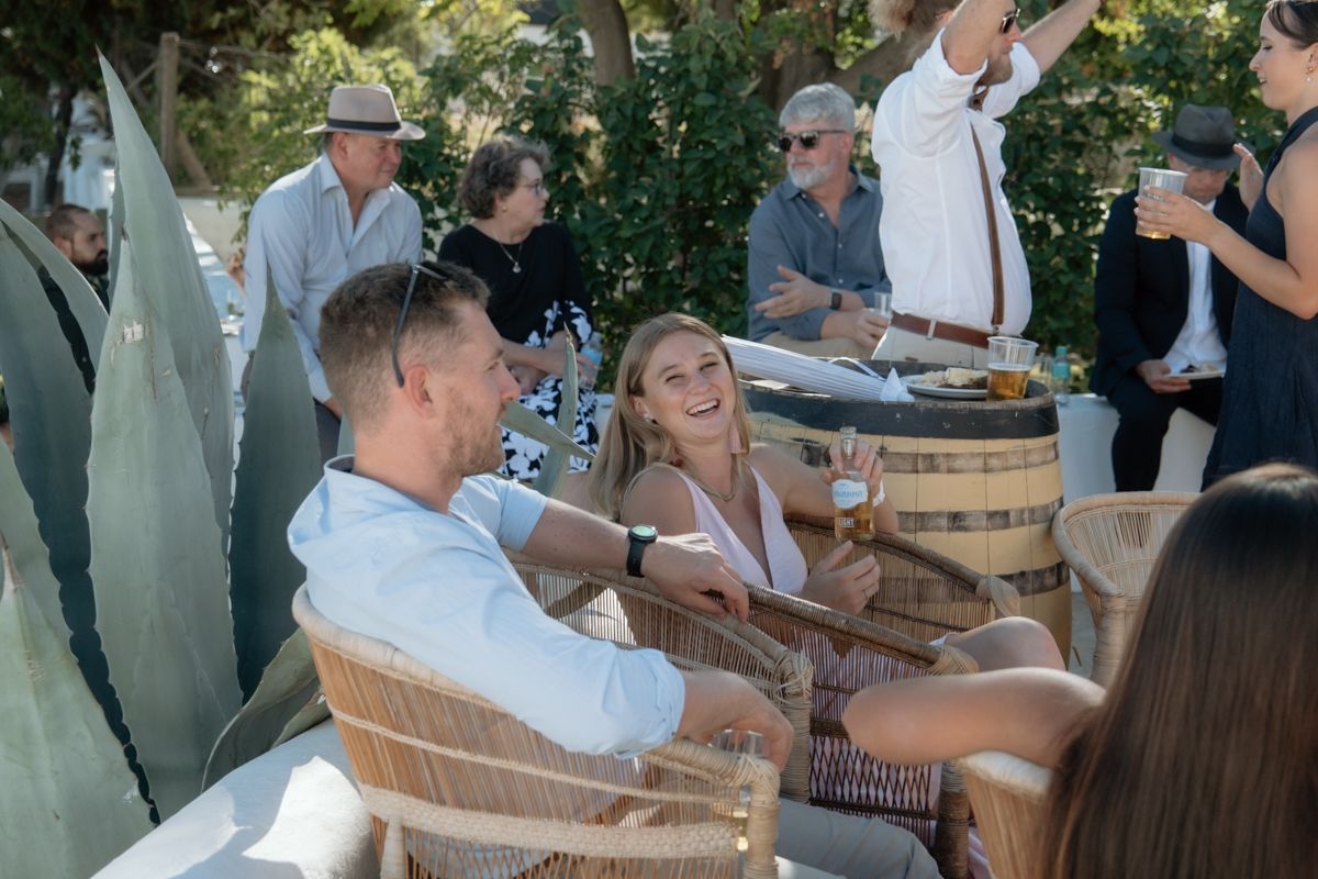 Couple laughing and enjoying drinks together at an outdoor wedding reception with guests in background.