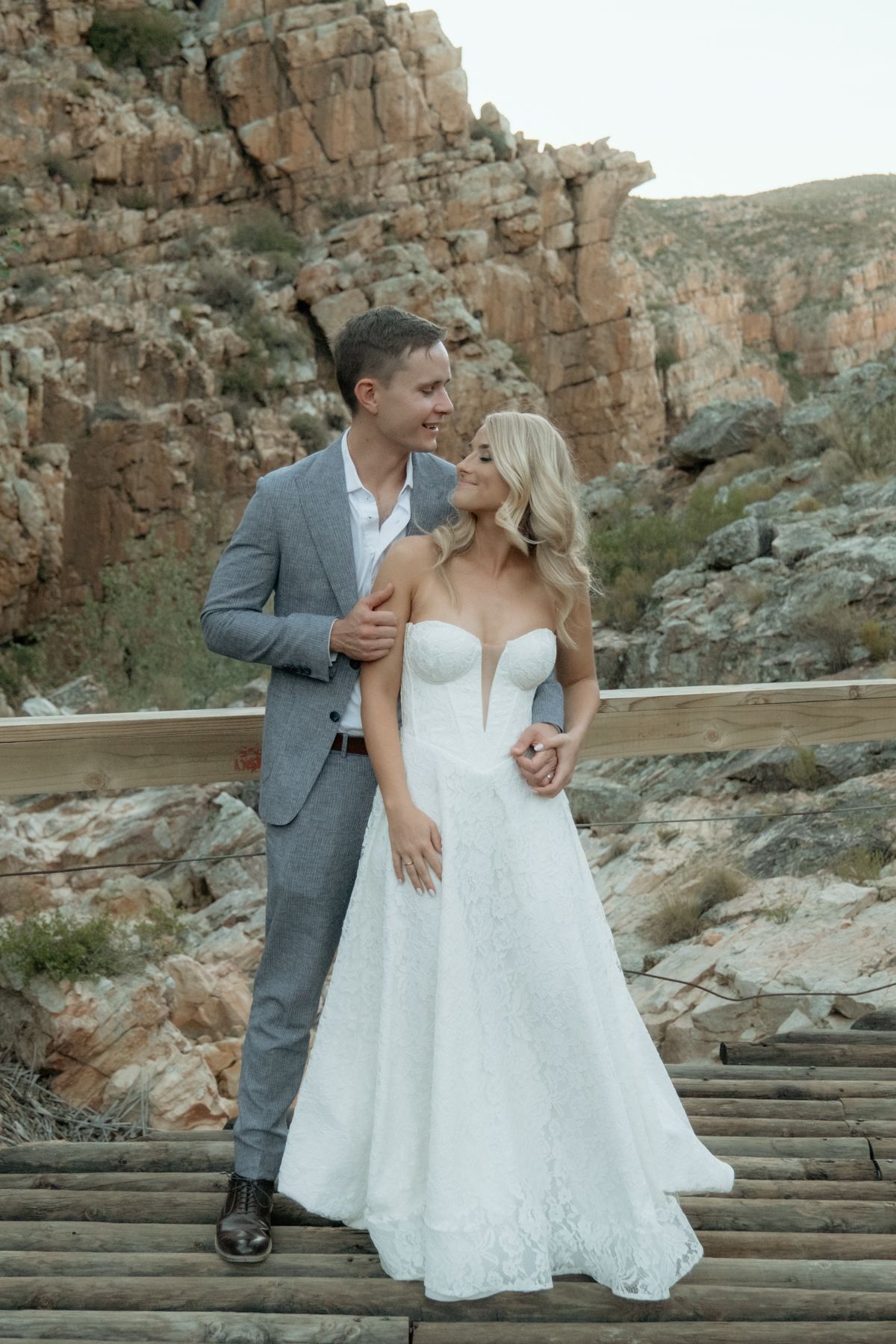 Bride and groom smiling at each other on wooden bridge with rocky canyon backdrop.