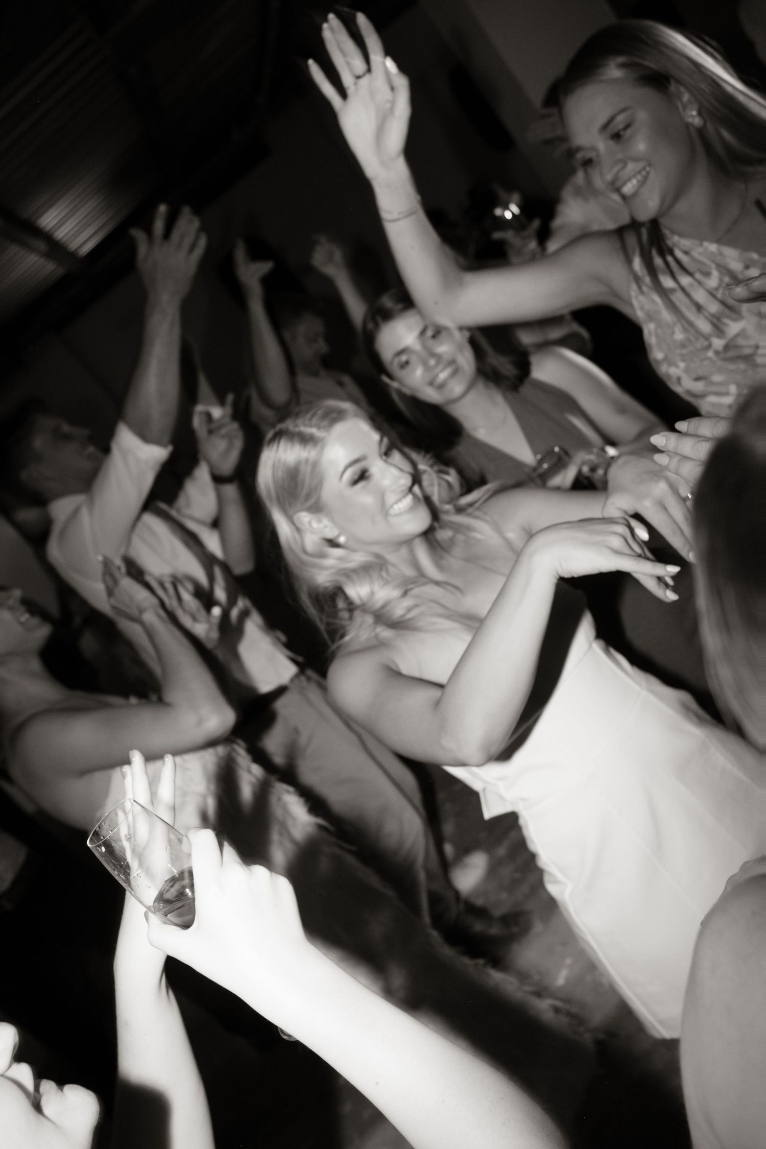 Black and white photo of bride dancing joyfully with wedding guests at lively indoor wedding reception celebration.