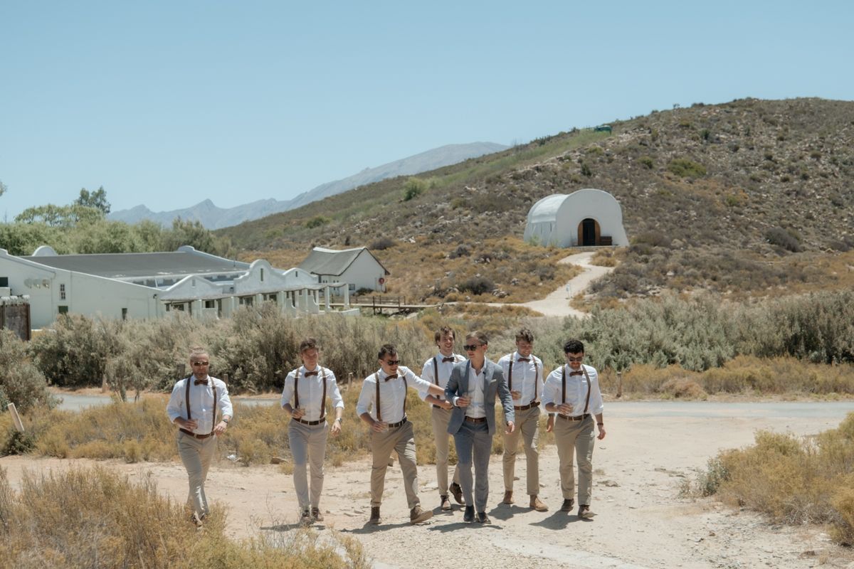 Groom and groomsmen in suspenders walking together on desert path with dome structure and mountains in background.