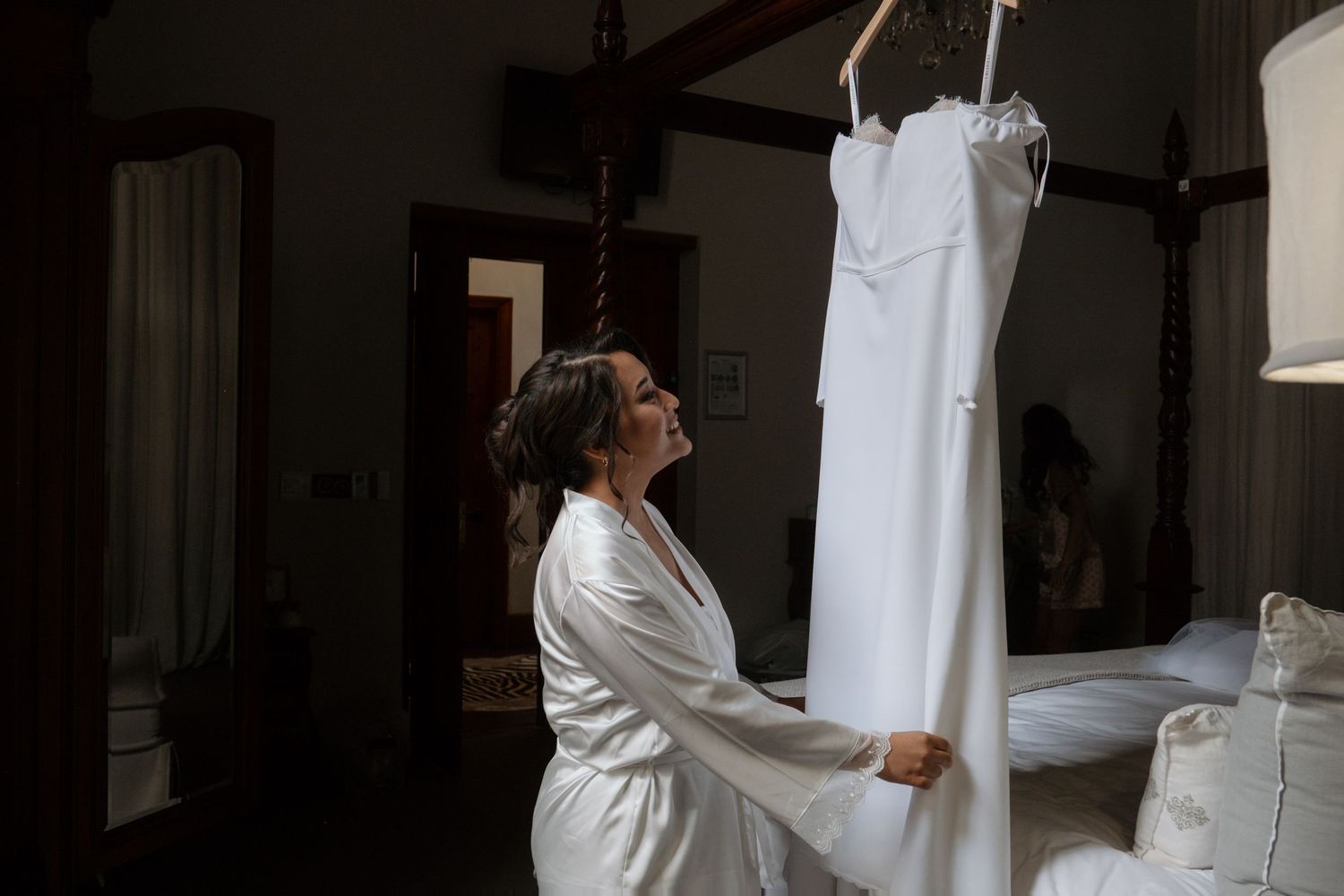 Bride in white robe admiring her wedding dress hanging in a elegantly decorated bedroom before the ceremony.