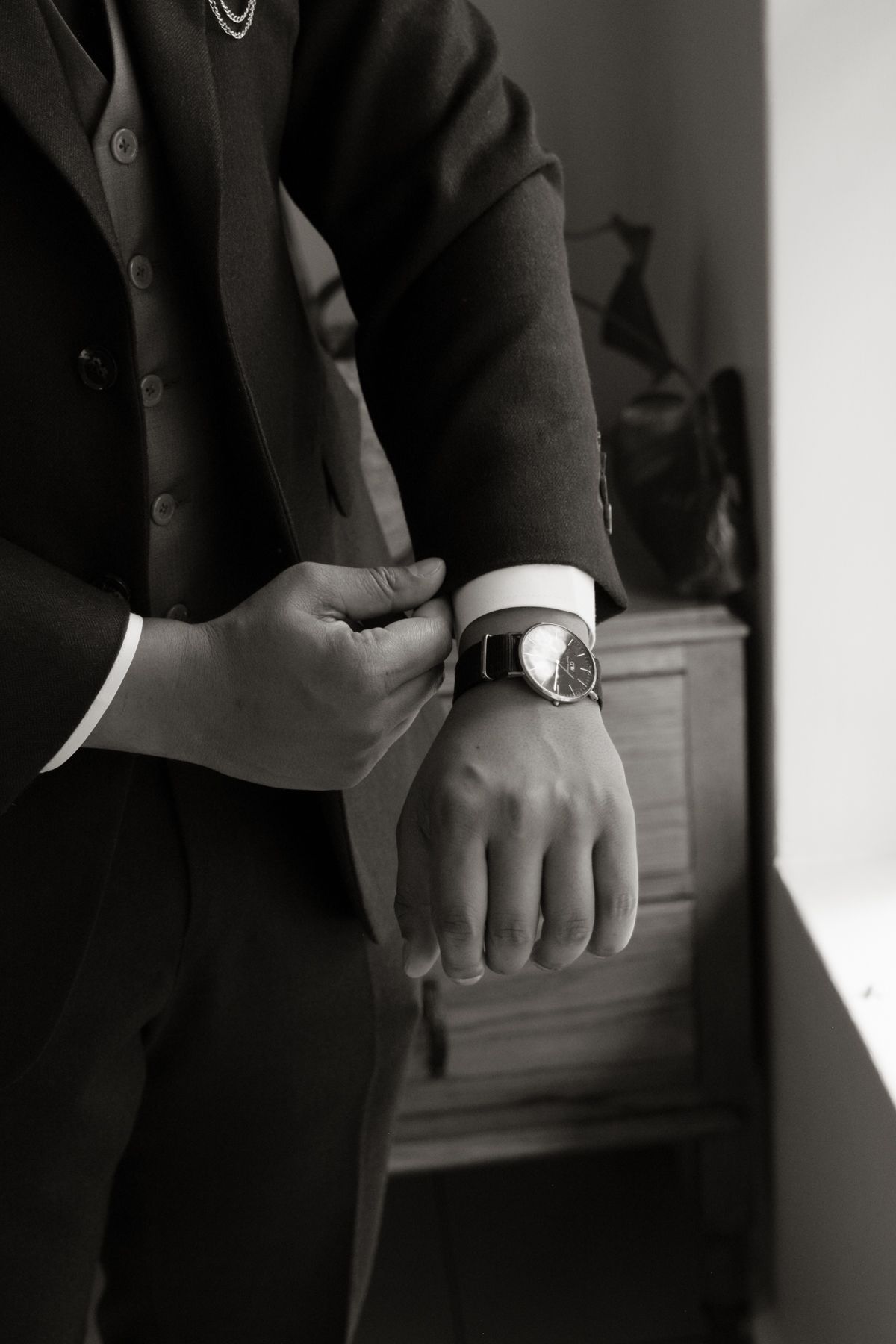 Black and white close-up of a groom in a dark suit adjusting his cufflinks and wristwatch before the ceremony.