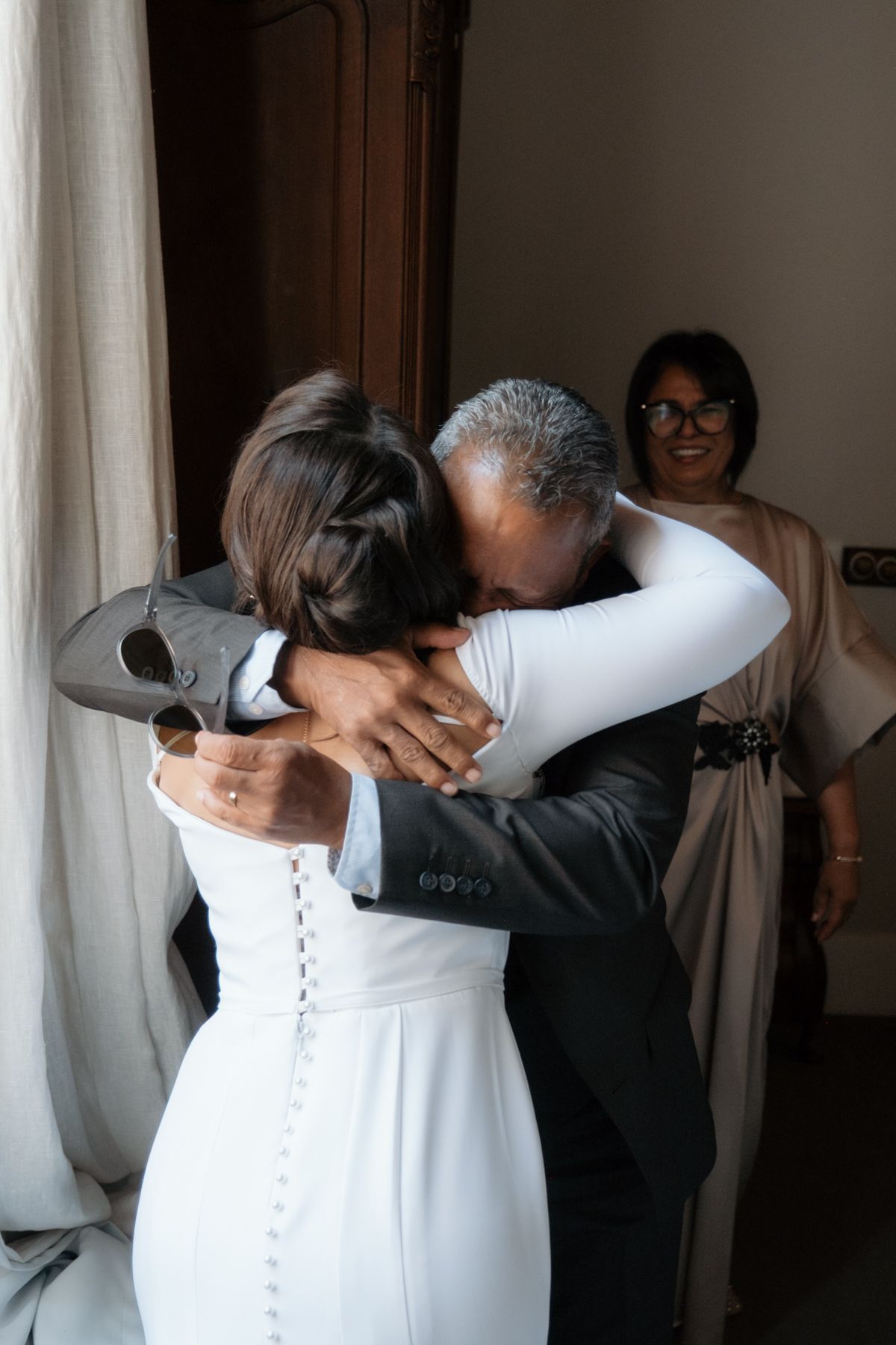 Bride in white gown shares emotional hug with father while smiling woman watches in background.