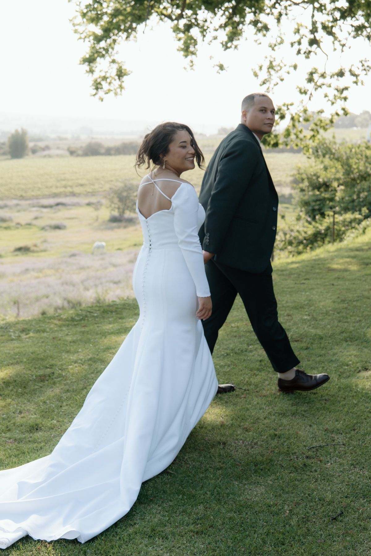 Bride in fitted white mermaid gown holds grooms hand while walking through lush green countryside landscape.