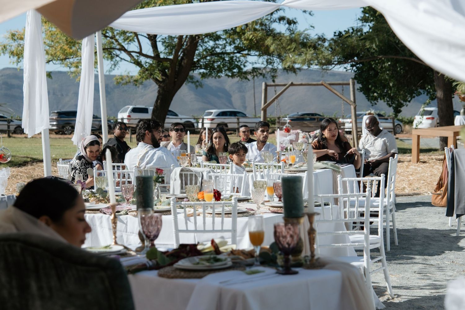 Outdoor wedding reception with guests seated at white-draped tables under a canopy, surrounded by trees and mountains.