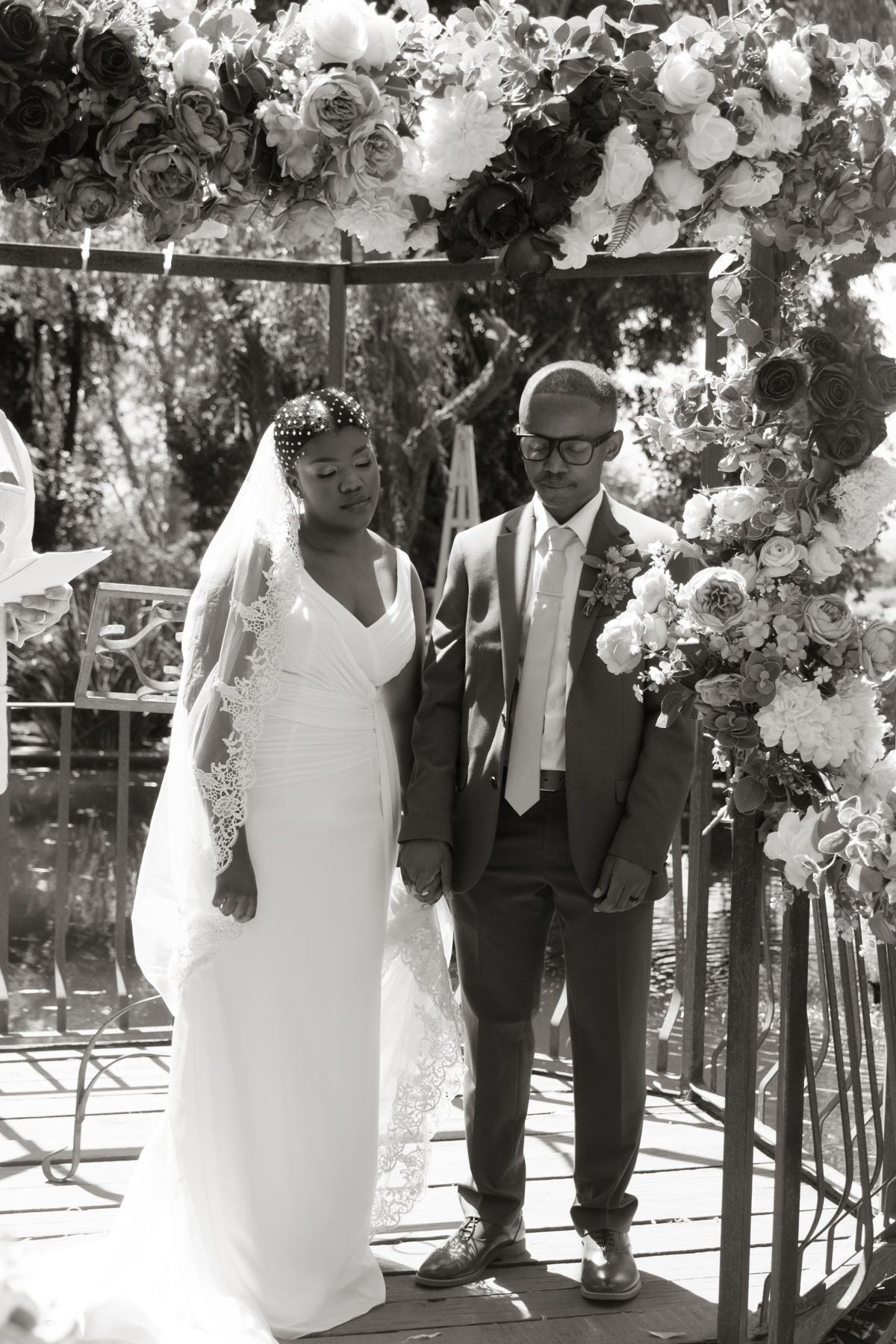 Black and white photo of bride and groom standing together under flower-adorned arch at wedding ceremony.