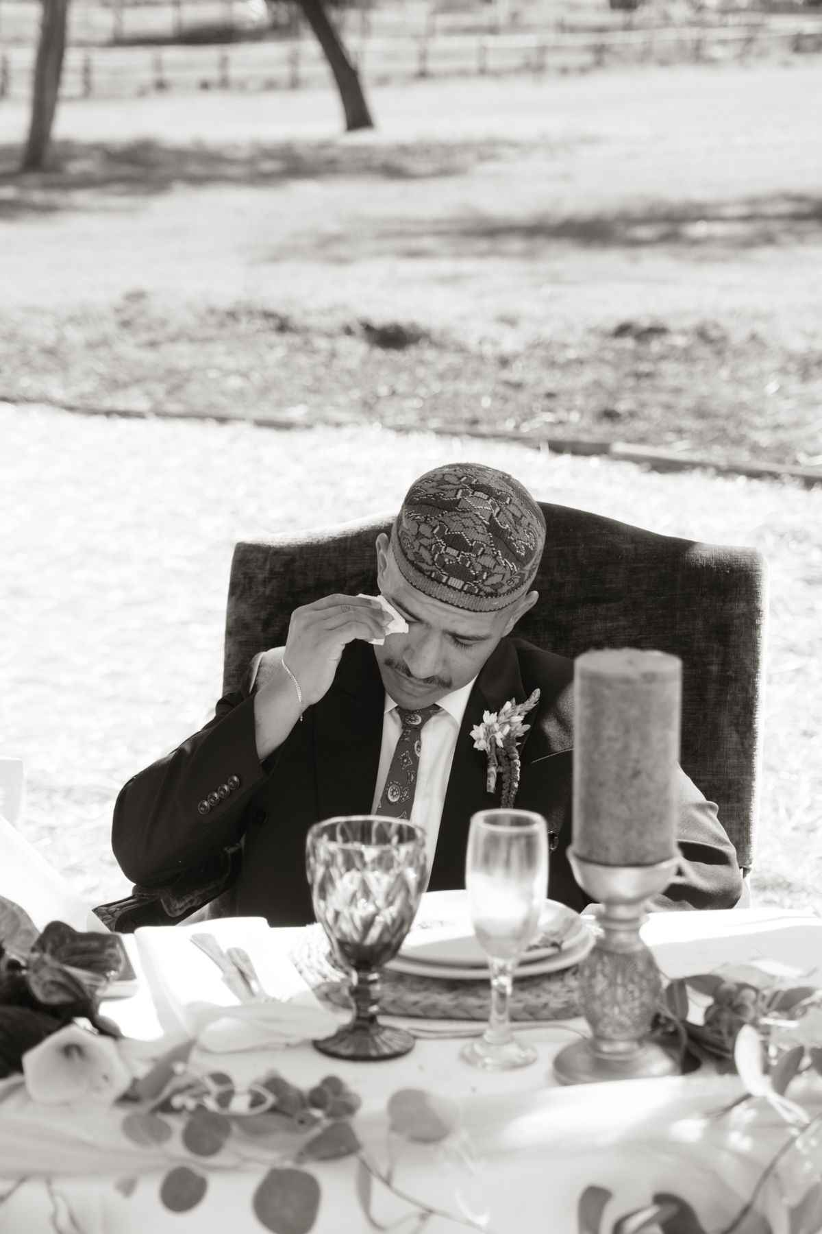 Black and white photo of groom in embroidered cap wiping tears at wedding reception table.