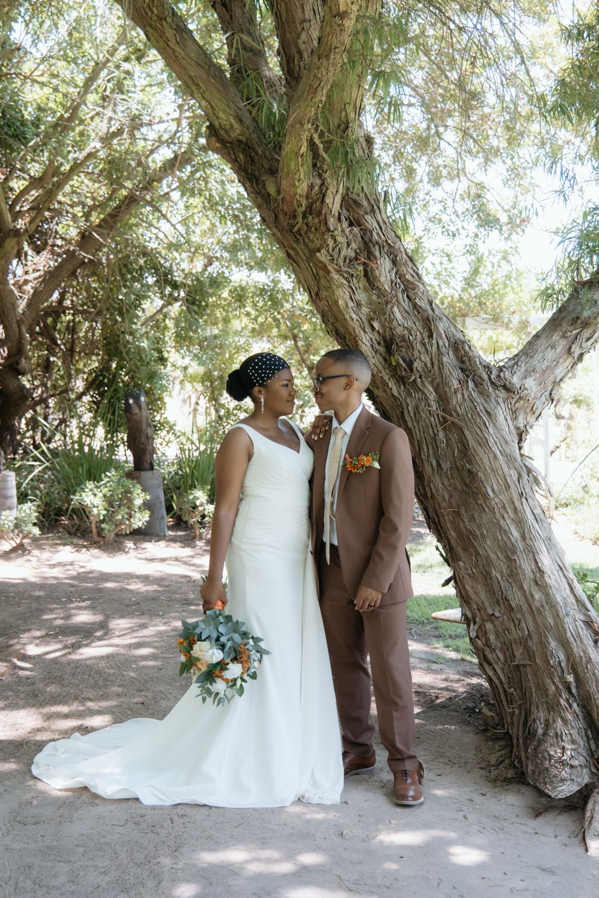 Black bride in white gown and groom in brown suit stand together under large tree holding floral bouquet.
