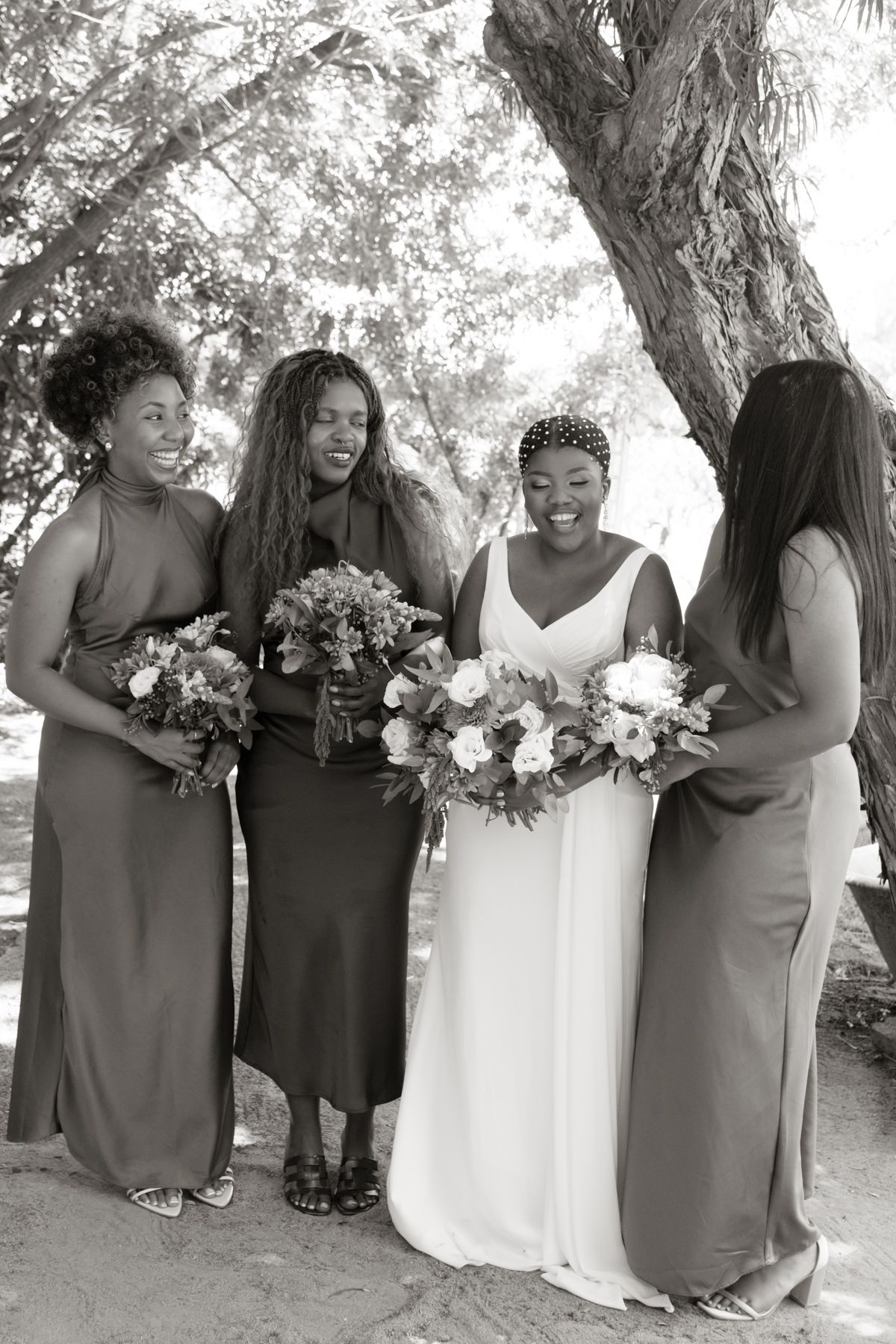 Black and white photo of bride and three bridesmaids holding bouquets under a tree outdoors.