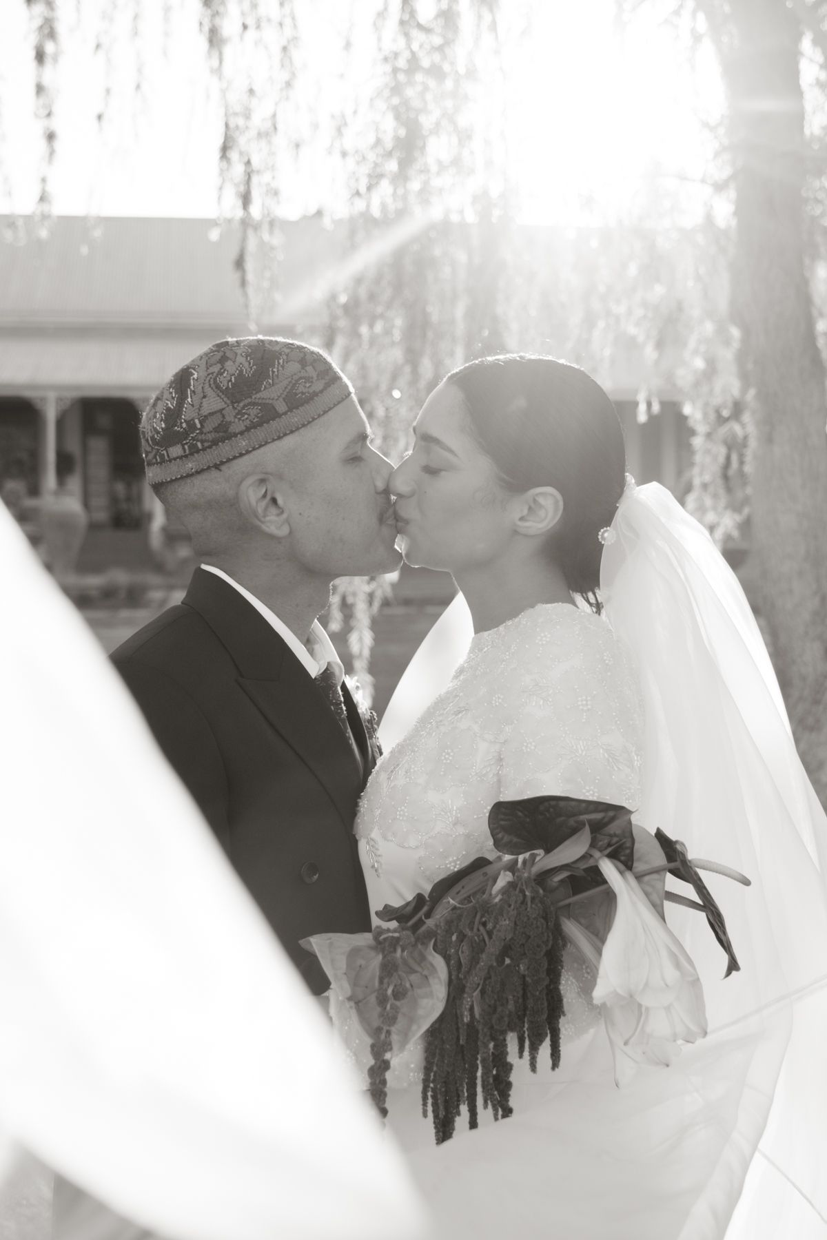 Black and white photo of interracial bride and groom sharing first kiss under weeping willow tree.
