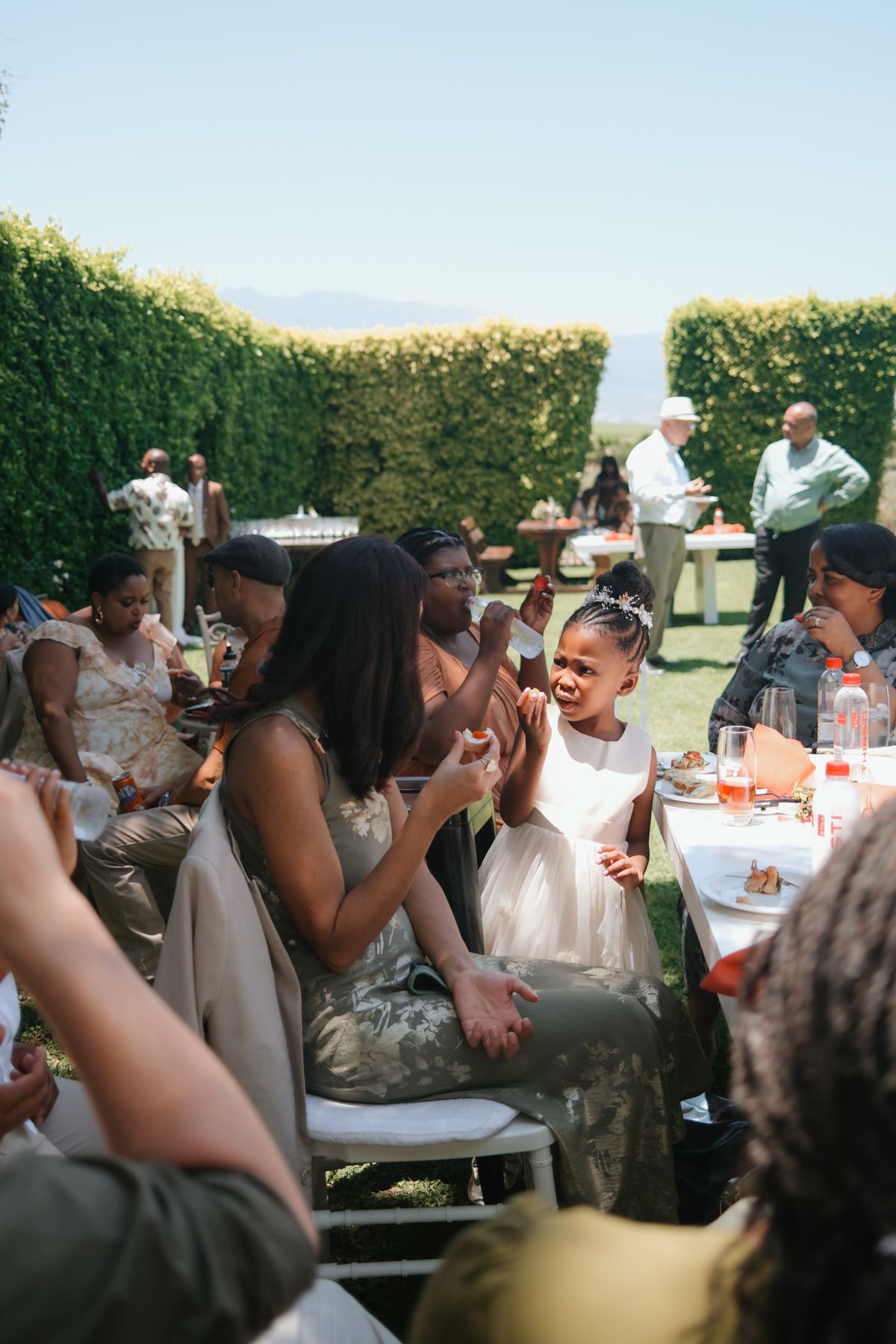 Joyful wedding guests clapping and celebrating at an outdoor garden reception with a young girl in a white dress.