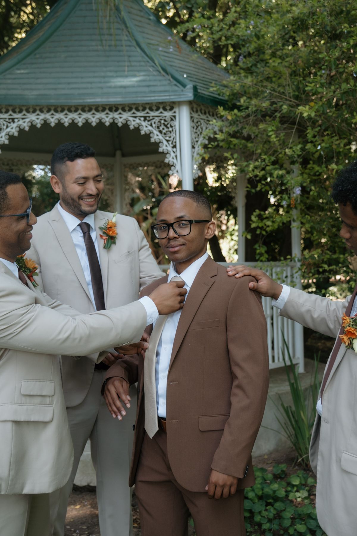 Groomsmen in tan suits help groom in brown suit adjust his tie near a gazebo outdoors.